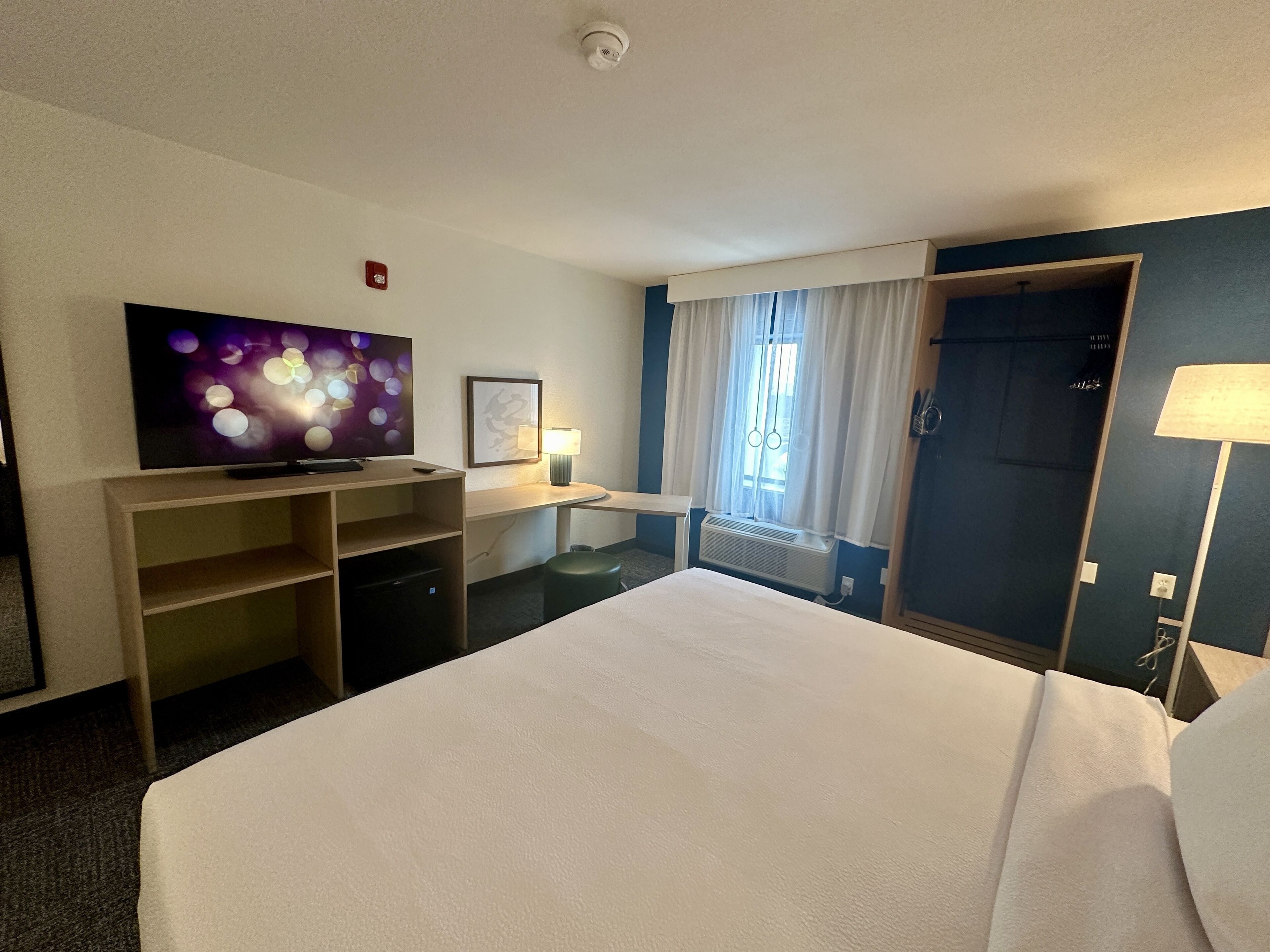 Habitación de hotel con cama, televisión, escritorio con lámpara, ventana con cortinas y puerta.