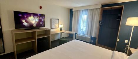 Habitación de hotel con cama, televisión, escritorio con lámpara, ventana con cortinas y puerta.