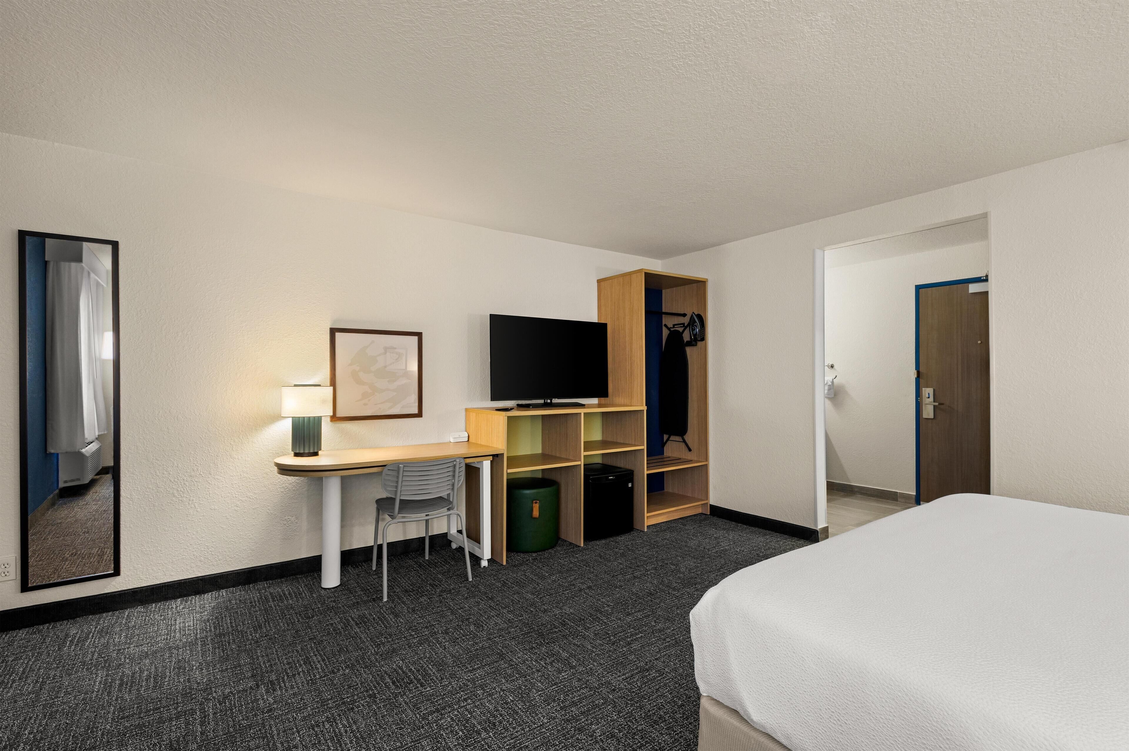 Habitación de hotel con cama, escritorio con silla, televisor, armario y espejo.