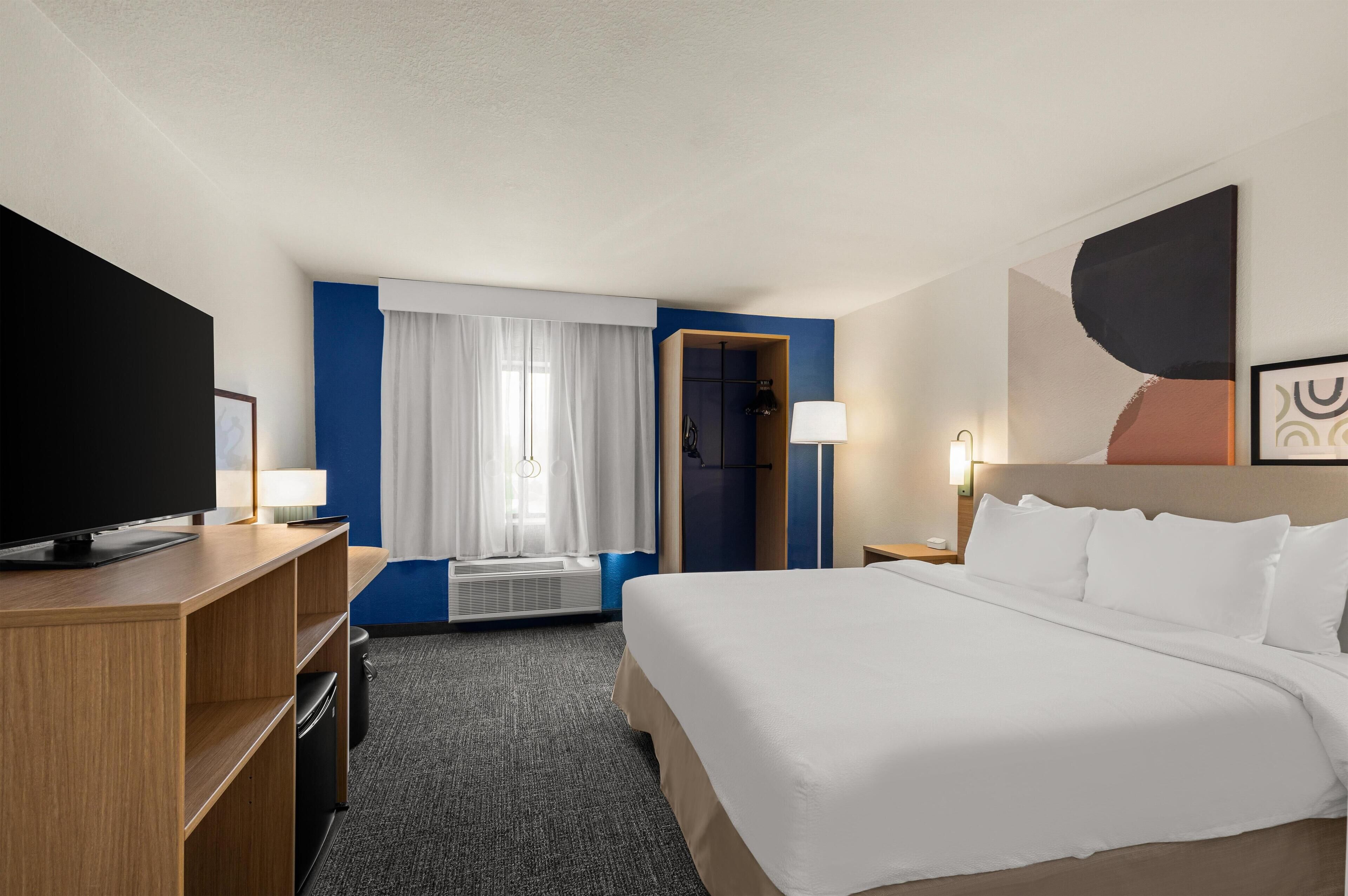 A hotel room with a large bed, a flat-screen TV, a desk with a lamp, and a window with curtains.