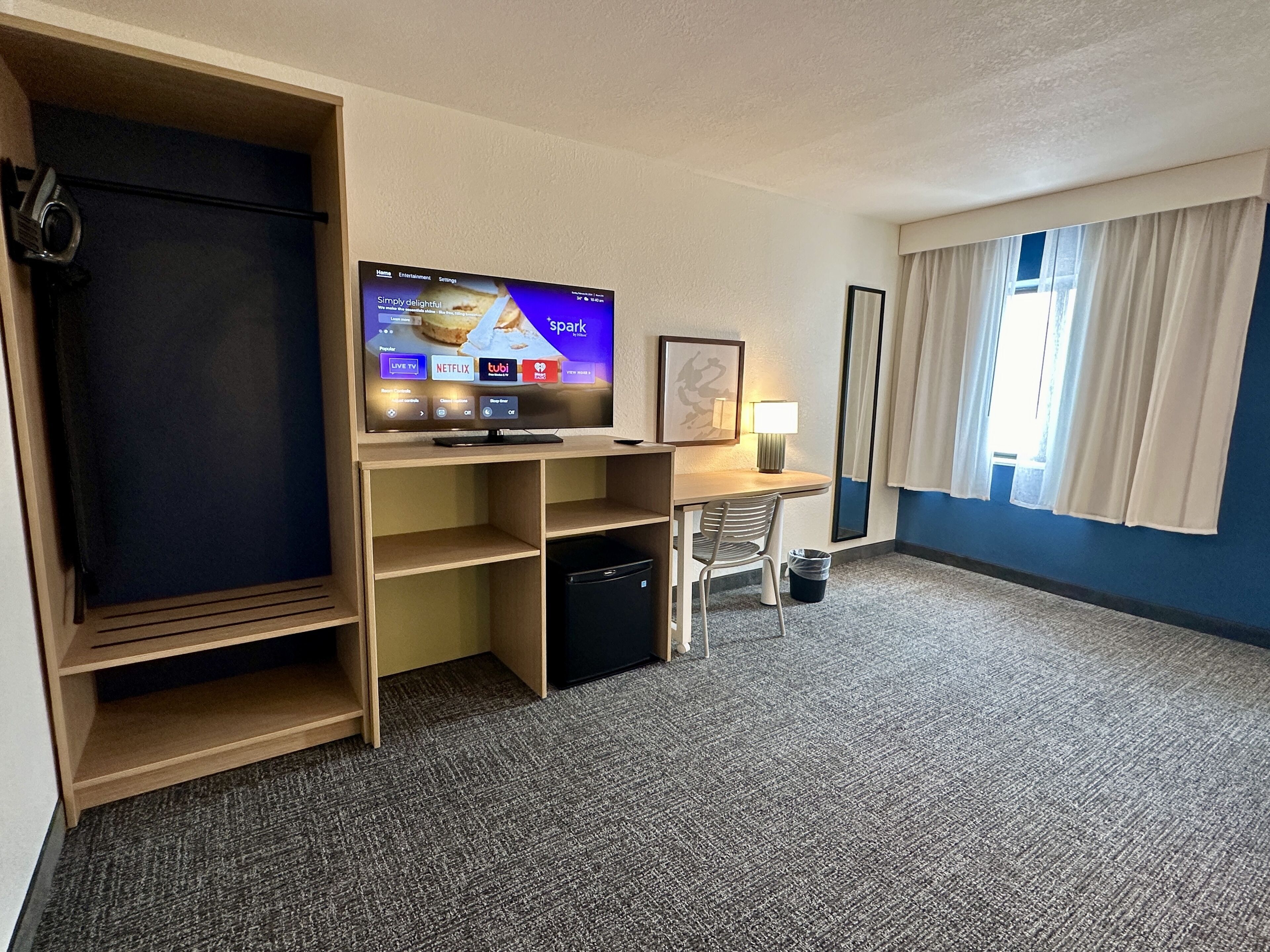 Une chambre d’hôtel avec un téléviseur posé sur un meuble en bois, un bureau avec une chaise, une lampe et une grande fenêtre avec des rideaux.