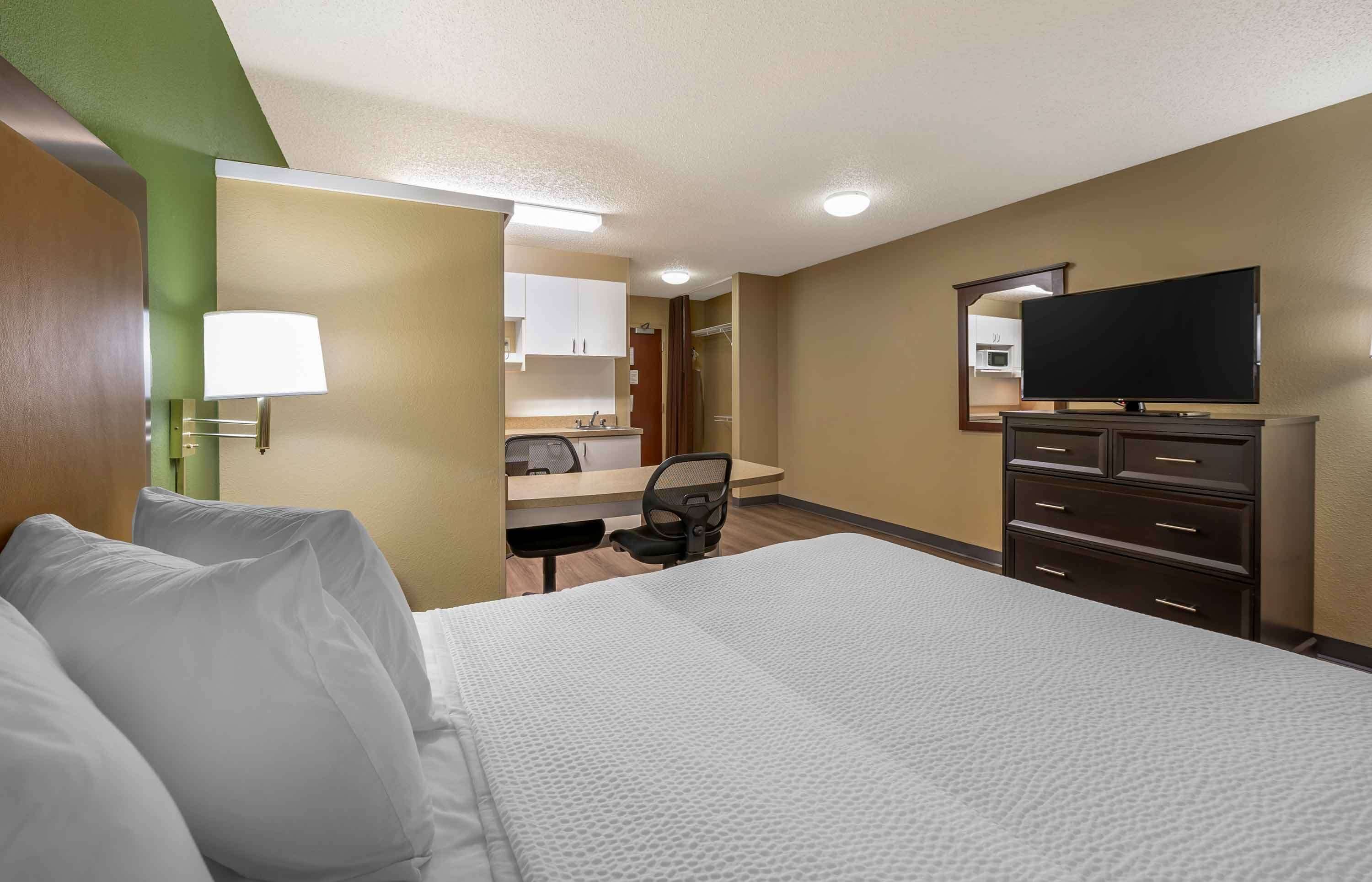 Quarto de hotel com cama, escrivaninha, cadeira, televisão e cômoda.