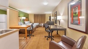 Ein Hotelzimmer mit Küchenzeile, einem Bett mit Kopfteil, einem Schreibtisch mit Stuhl, einem Fernseher und einem gerahmten Bild an der Wand.