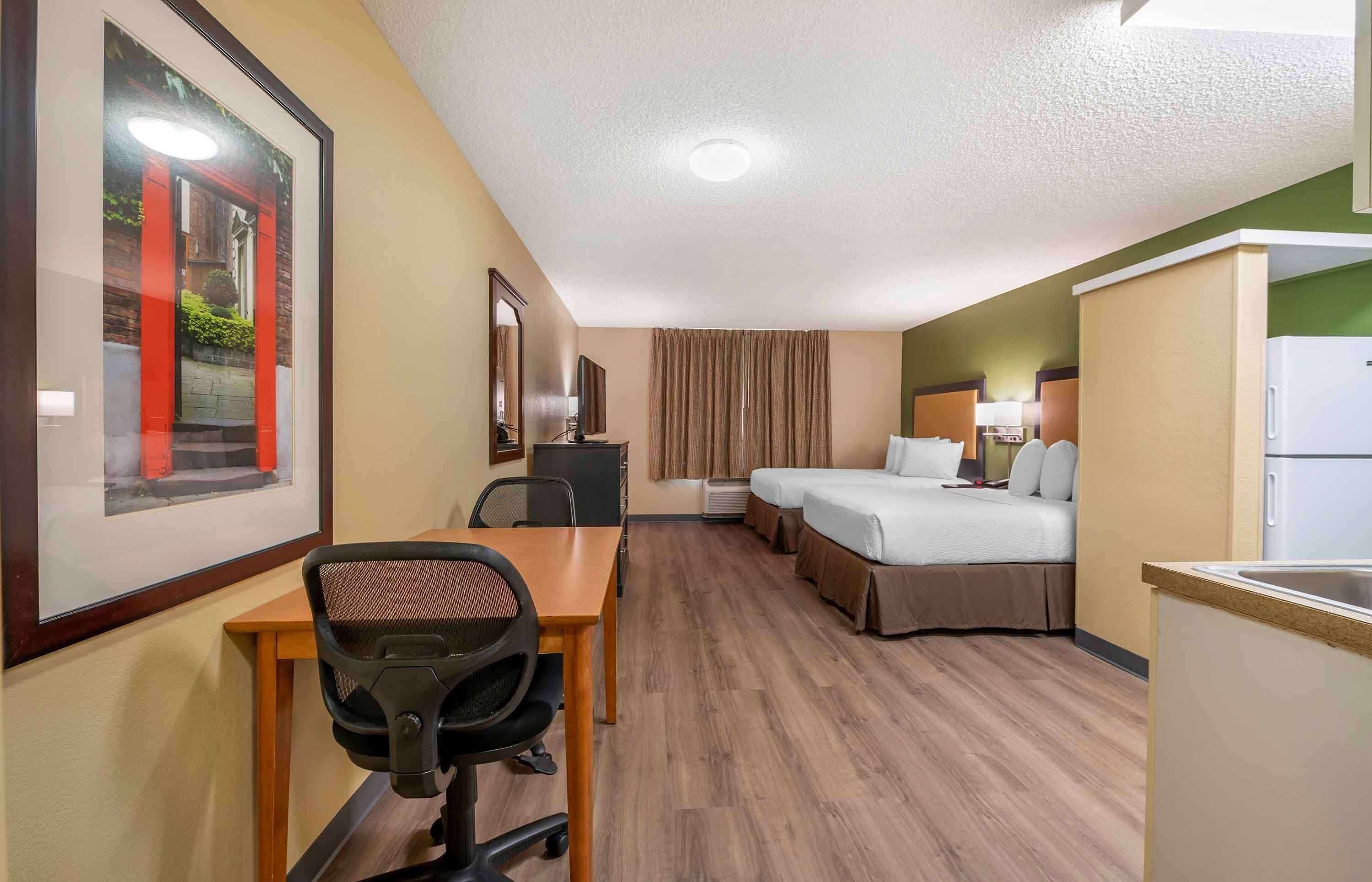 A hotel room with a wooden desk, two beds, a kitchenette, and a framed picture on the wall.