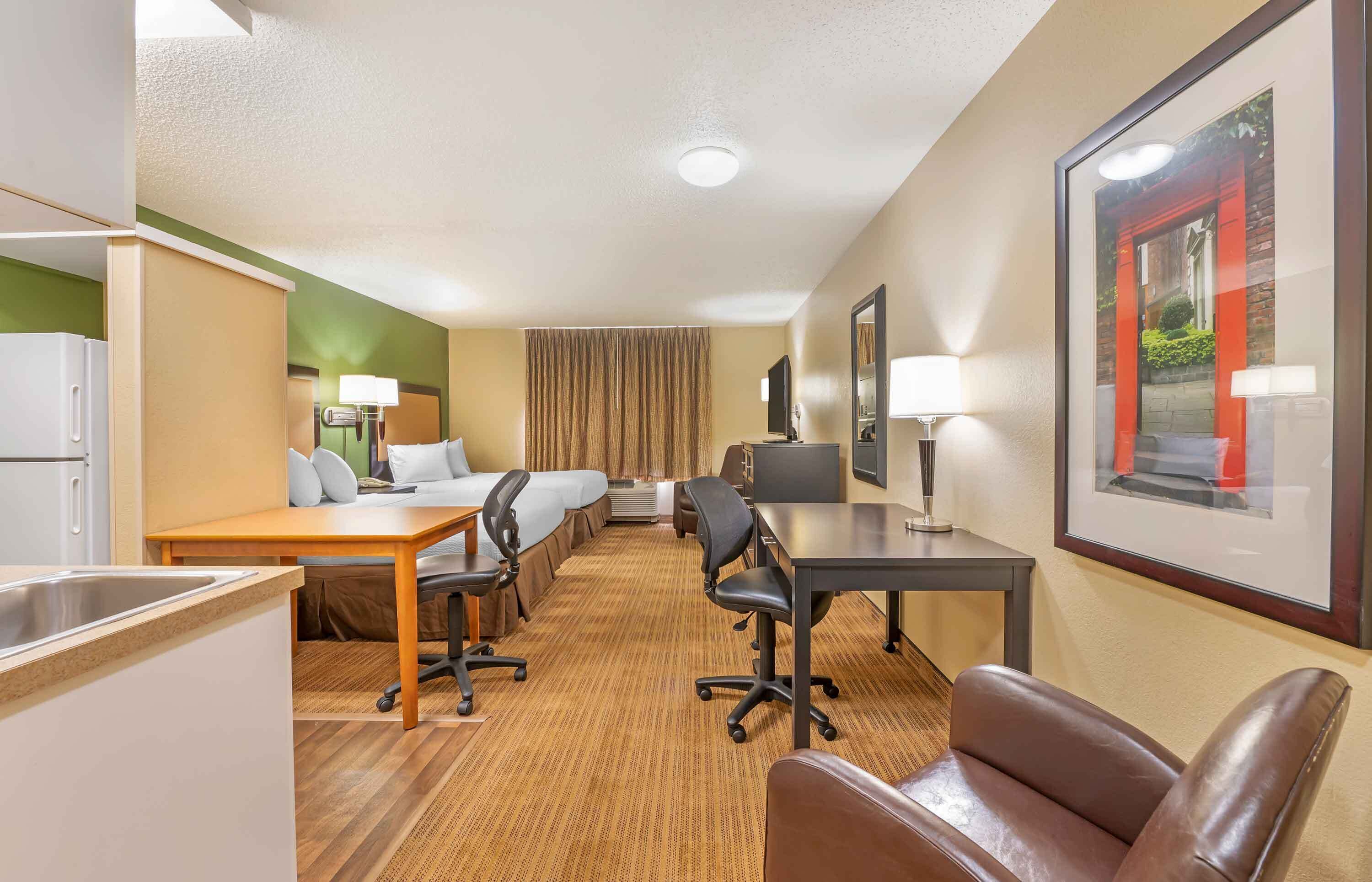 A hotel room with a kitchenette, a dining area with a table and chairs, a bed with a headboard, a desk with a chair, a large window with curtains, and a framed picture on the wall.