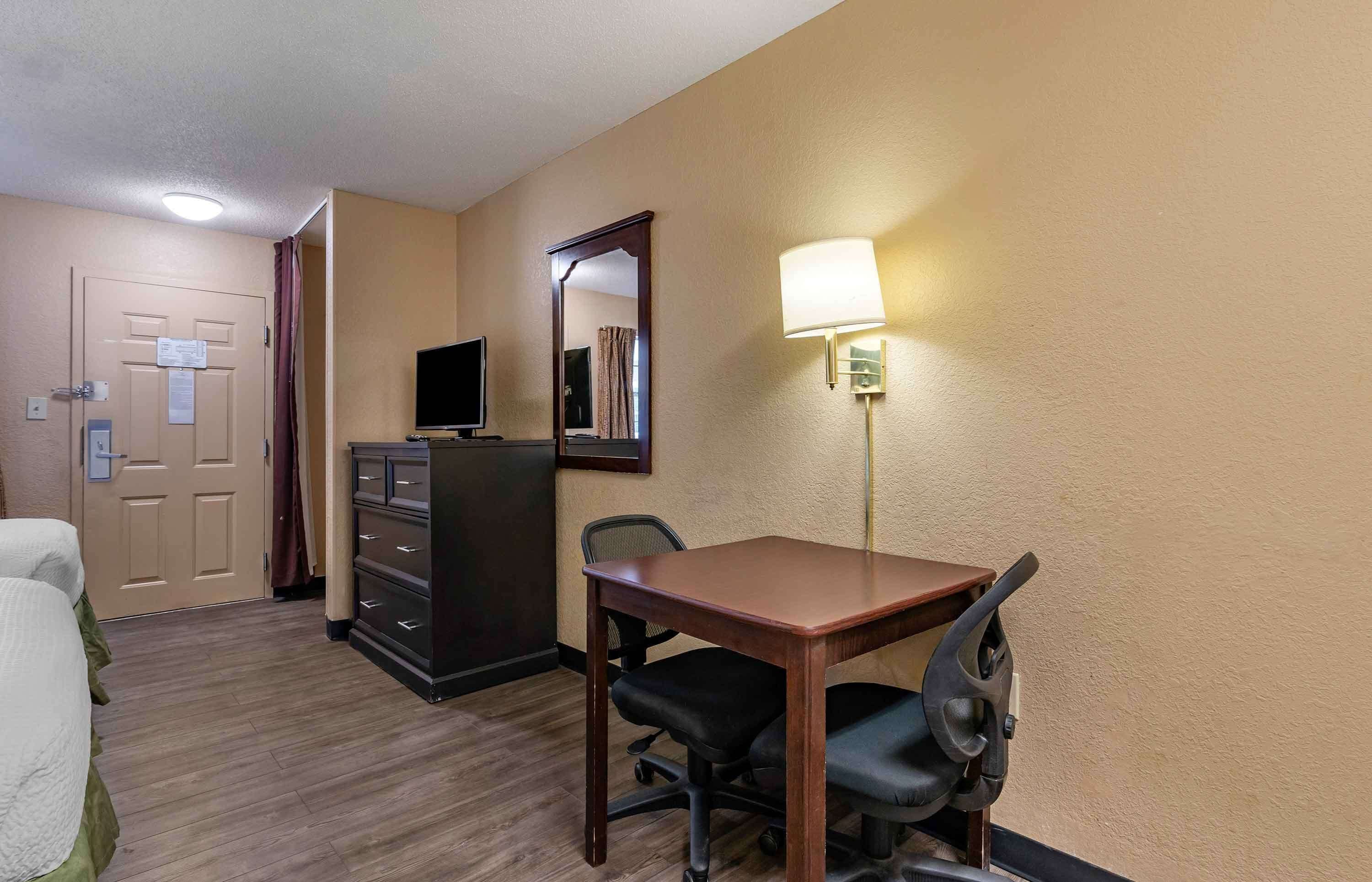 A hotel room with a wooden desk, two chairs, a dresser, a television, and a lamp.