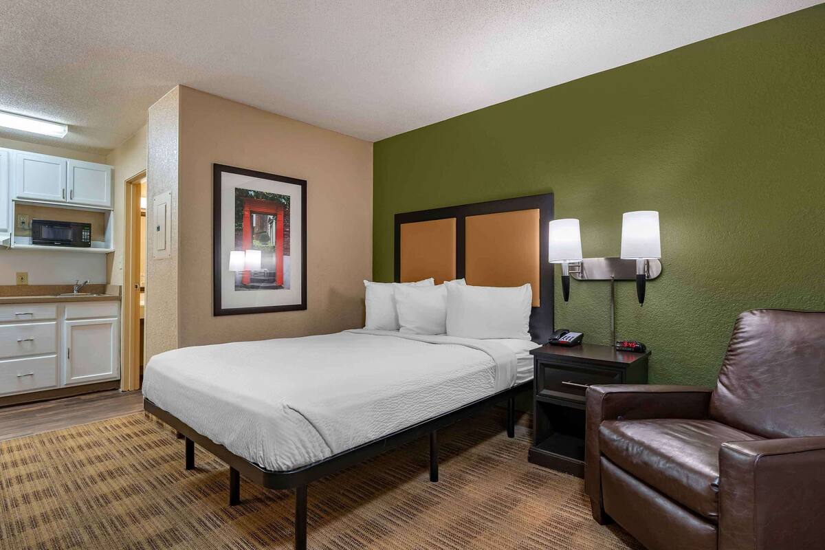 Una camera d'hotel con un letto, una poltrona in pelle marrone, un comodino con una lampada, un forno a microonde e un quadro appeso al muro.