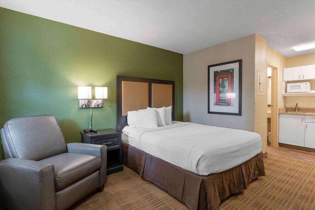 Camera d'albergo con un letto, una sedia, un comodino, una lampada, un quadro incorniciato e un angolo cottura.