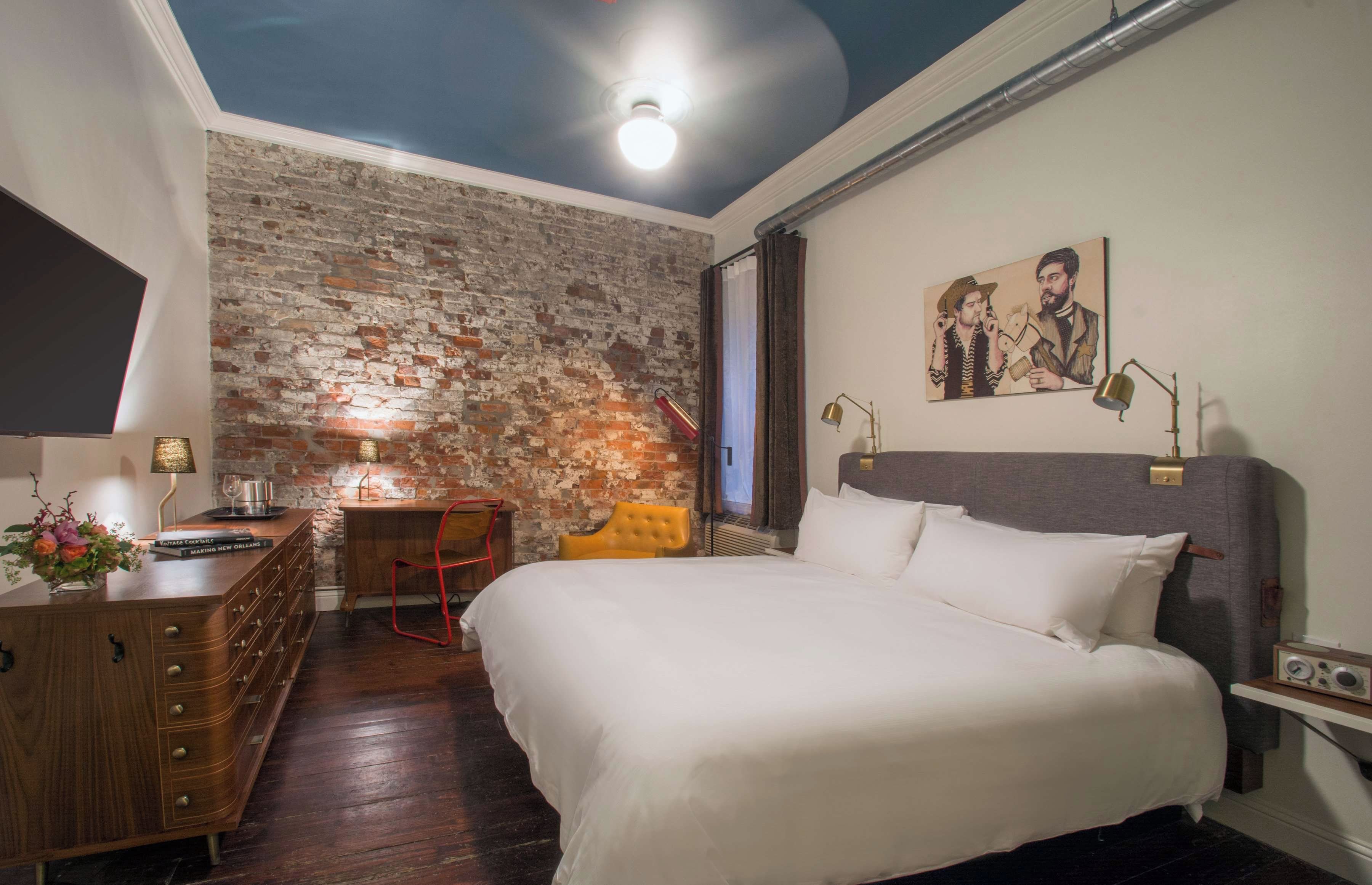 A hotel room with a brick wall, a bed, a desk with a lamp, a chair, and a painting on the wall.