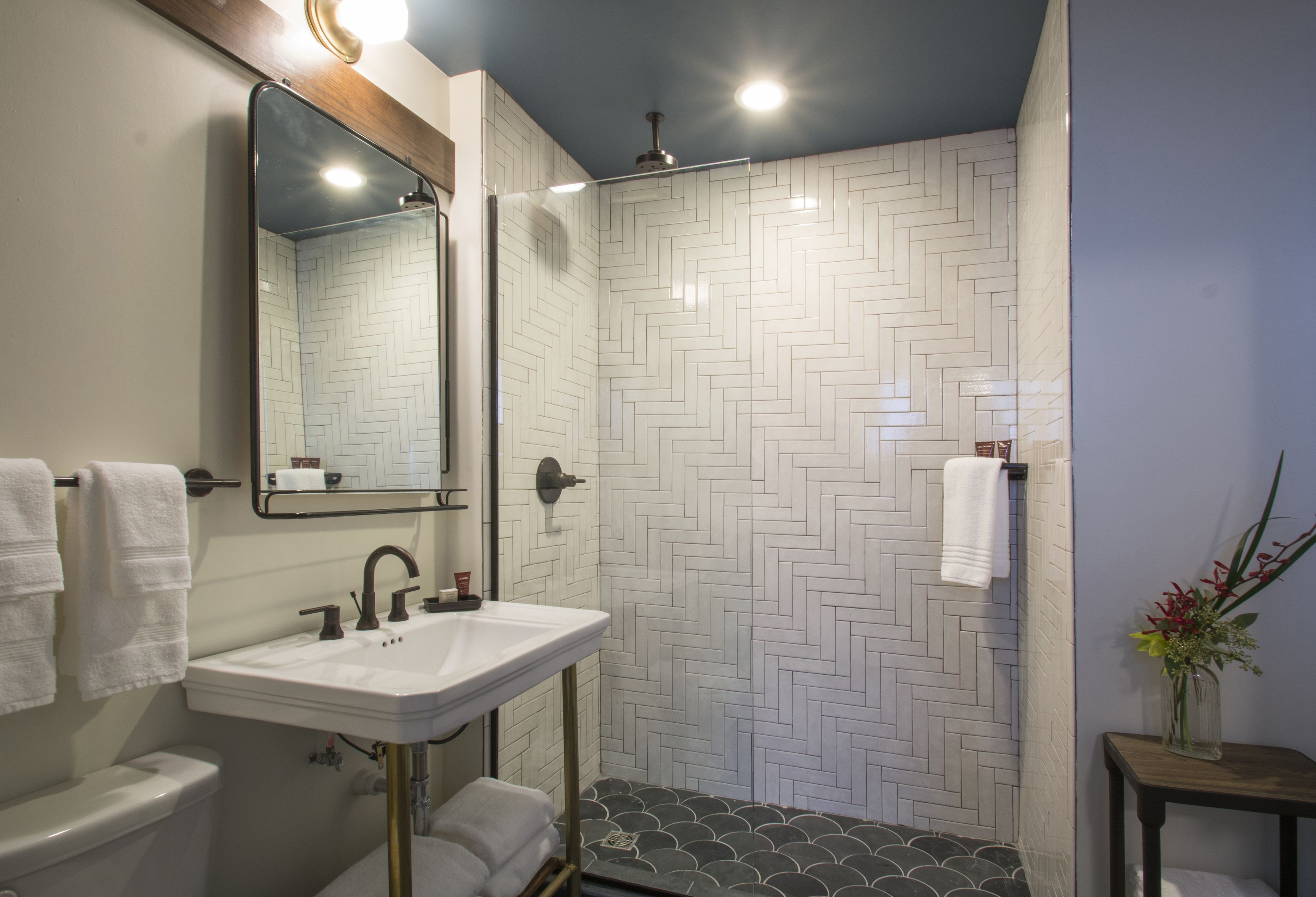 Une salle de bain avec un carrelage blanc disposé en motif de chevron, un lavabo blanc, un miroir et une douche.