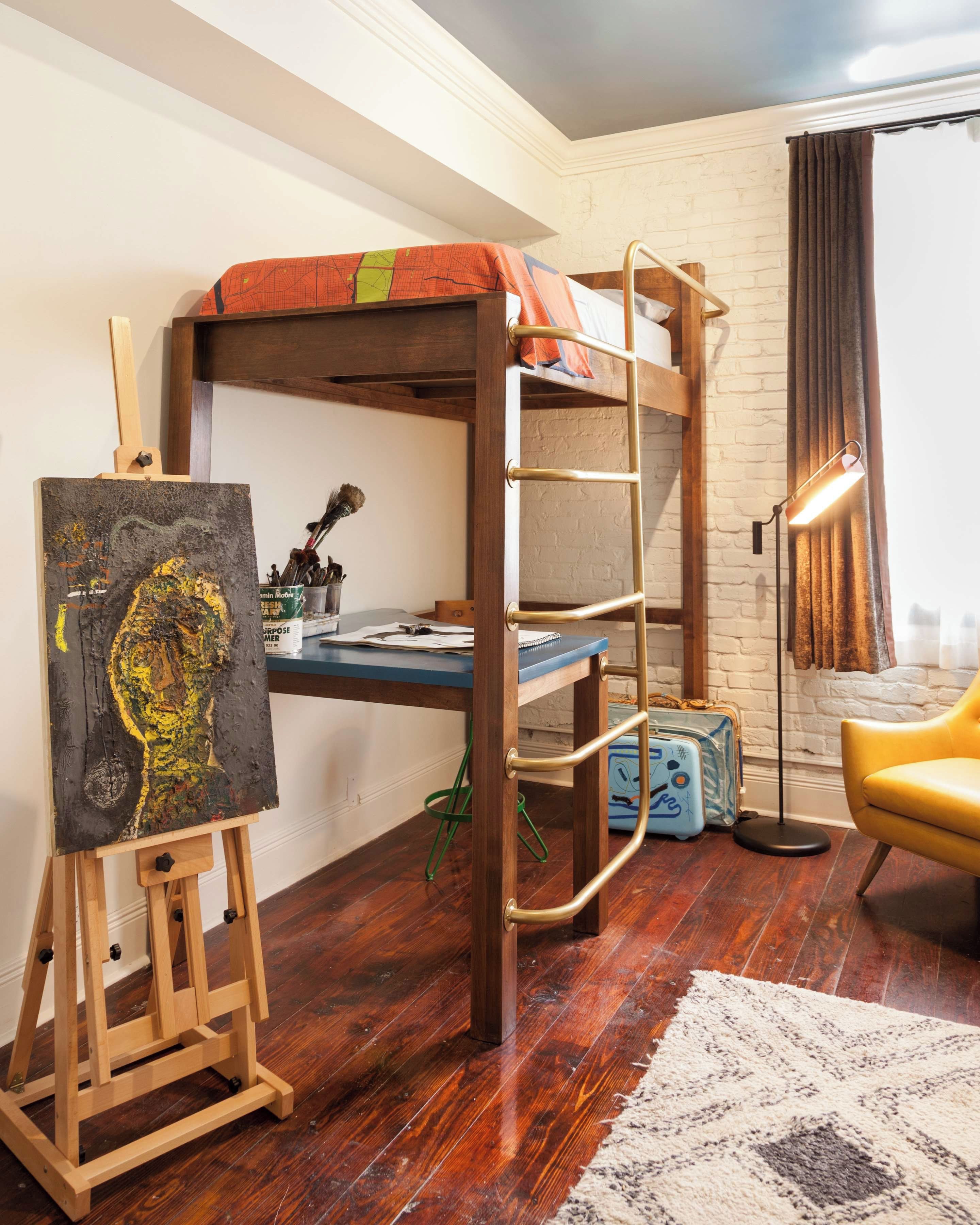 A bunk bed with a desk, an easel, and a lamp in a room with a brick wall and a window with curtains.