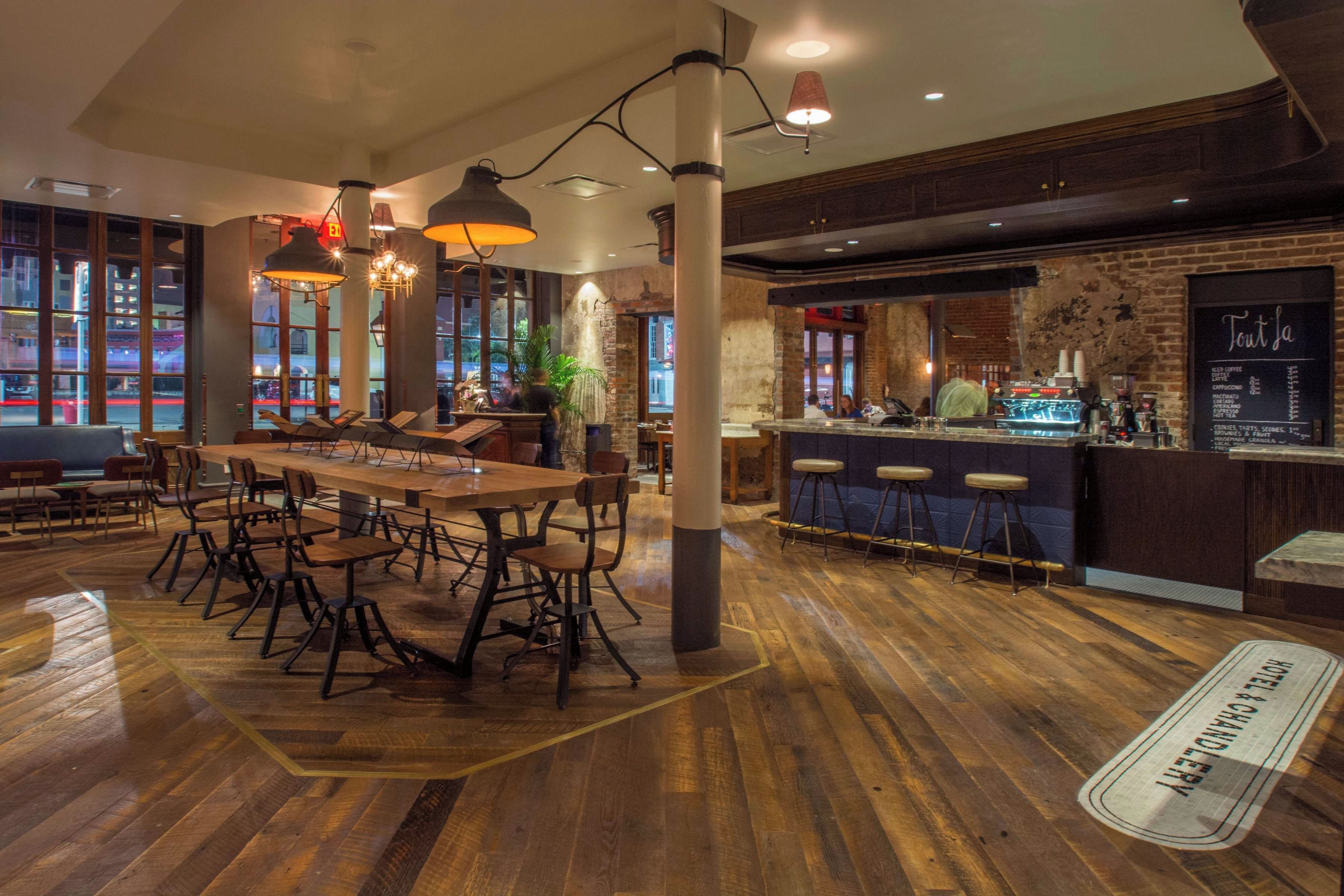 A restaurant interior with a long wooden table, multiple chairs, hanging pendant lights, and a bar area with stools.