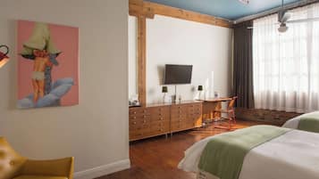 Ein Hotelzimmer mit einem Holzkoffer, einem Fernseher, einem Schreibtisch, zwei Betten, einem Sessel und einem Bild an der Wand.