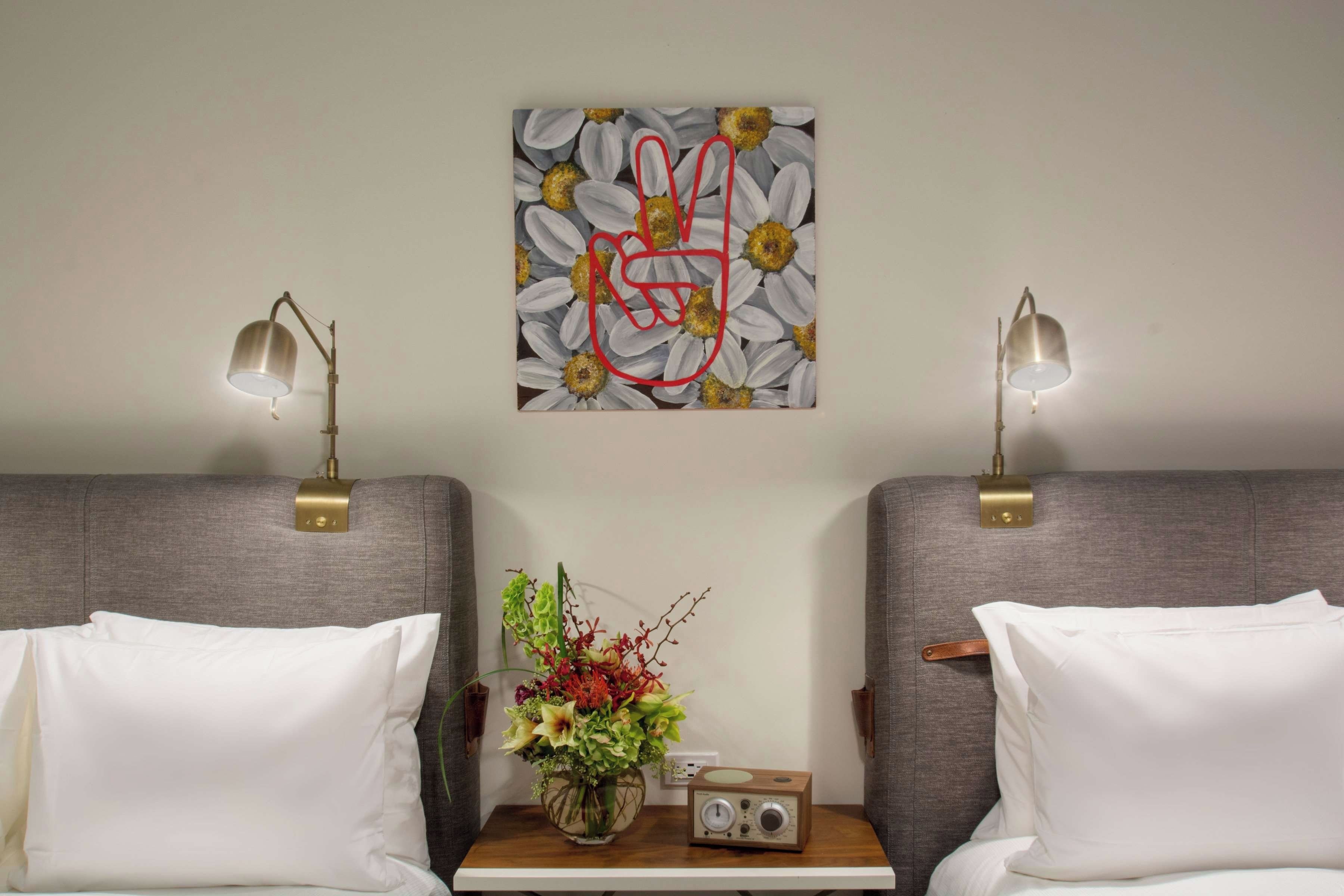 A hotel room with two beds, a nightstand with a radio and flowers, and a wall art piece above the beds.