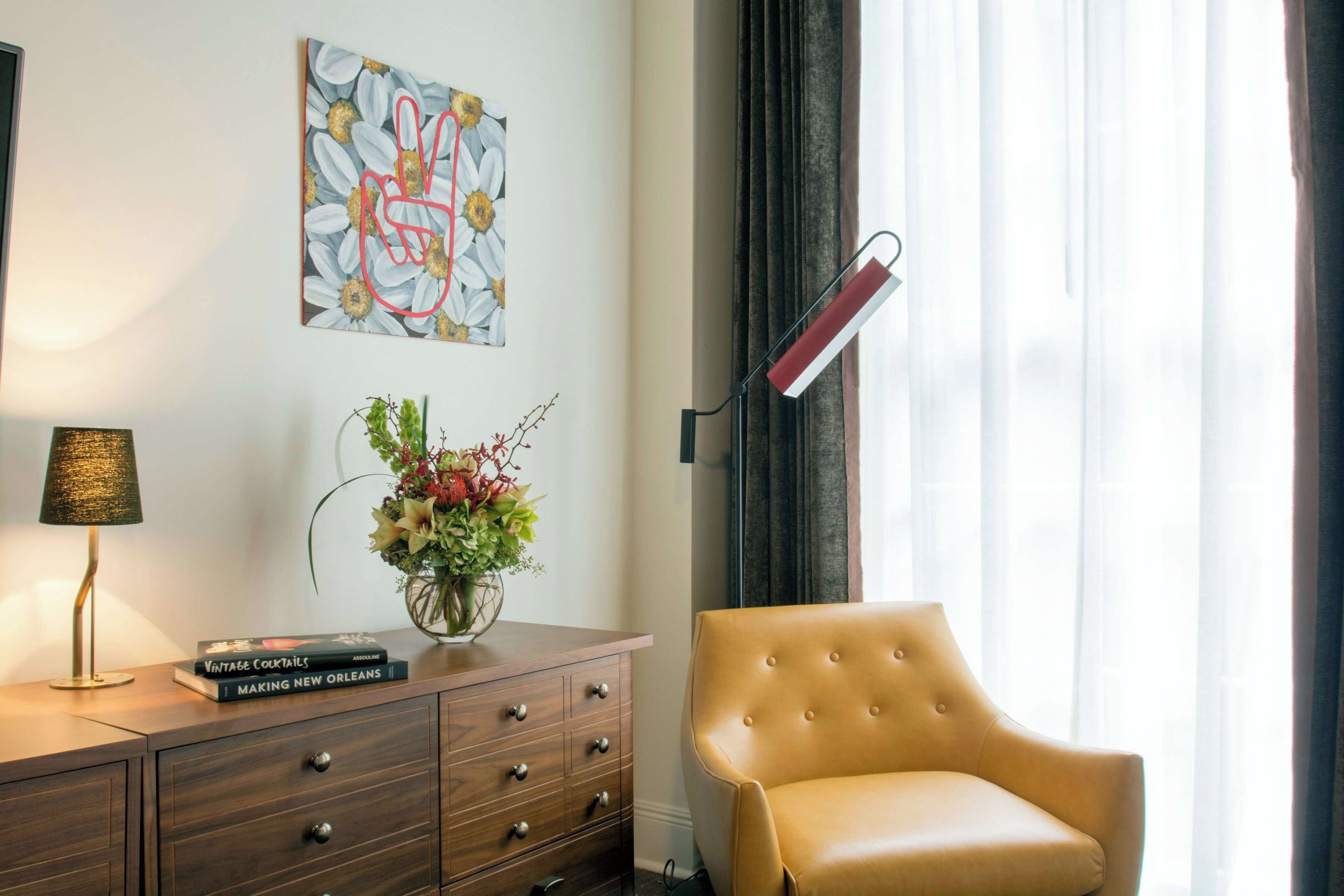 Una habitación de hotel con un aparador de madera, un florero con flores, un libro titulado 'Making New Orleans', una lámpara, una silla y un cuadro en la pared.