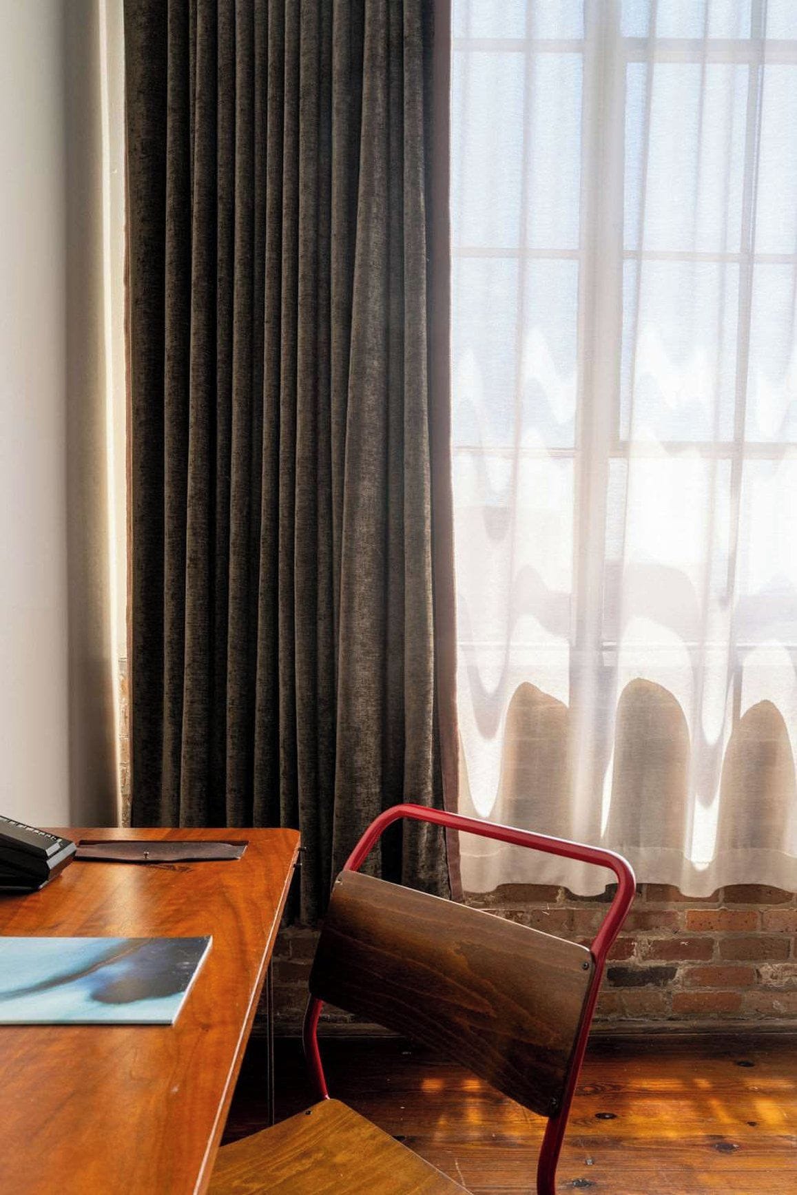 A room with a wooden desk, a red metal chair, a window with sheer curtains, and a brick wall.