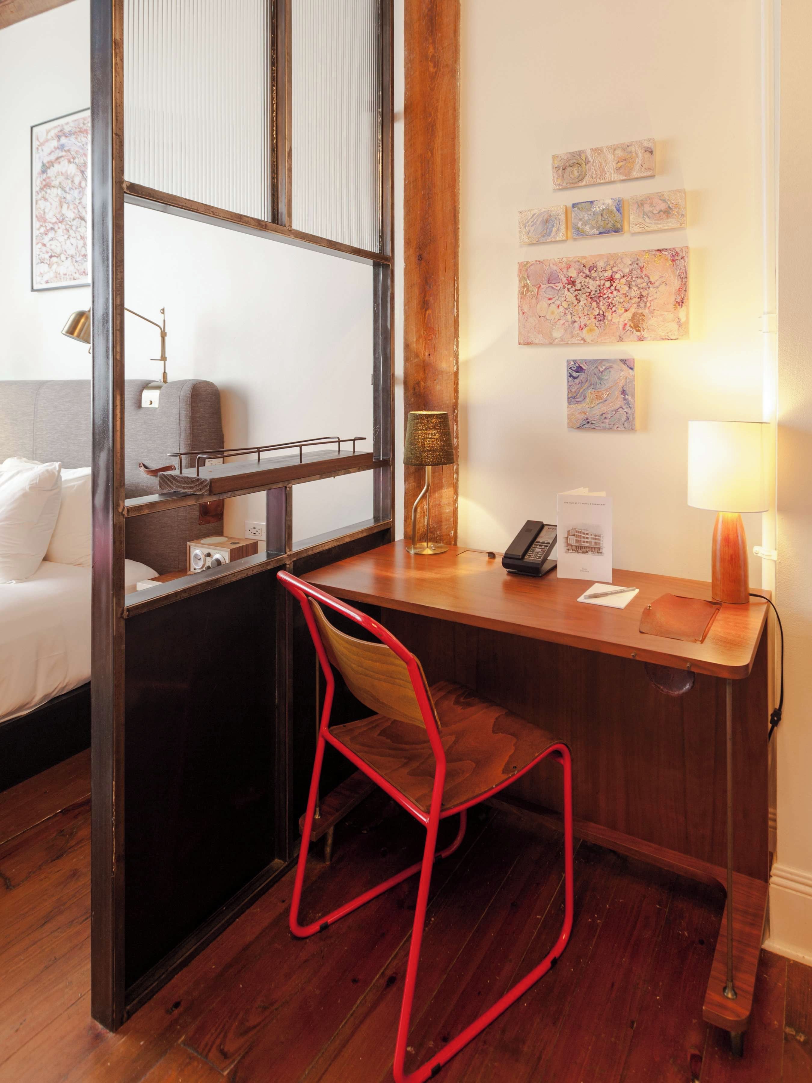 A hotel room with a wooden desk, a red chair, a bed with white linens, a wall-mounted lamp, and artwork.