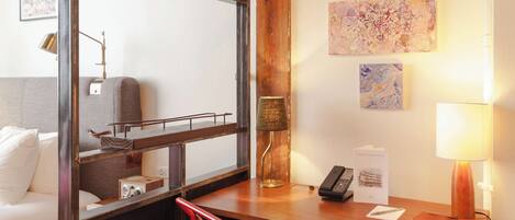 A hotel room with a wooden desk, a red chair, a bed with white linens, a wall-mounted lamp, and artwork.