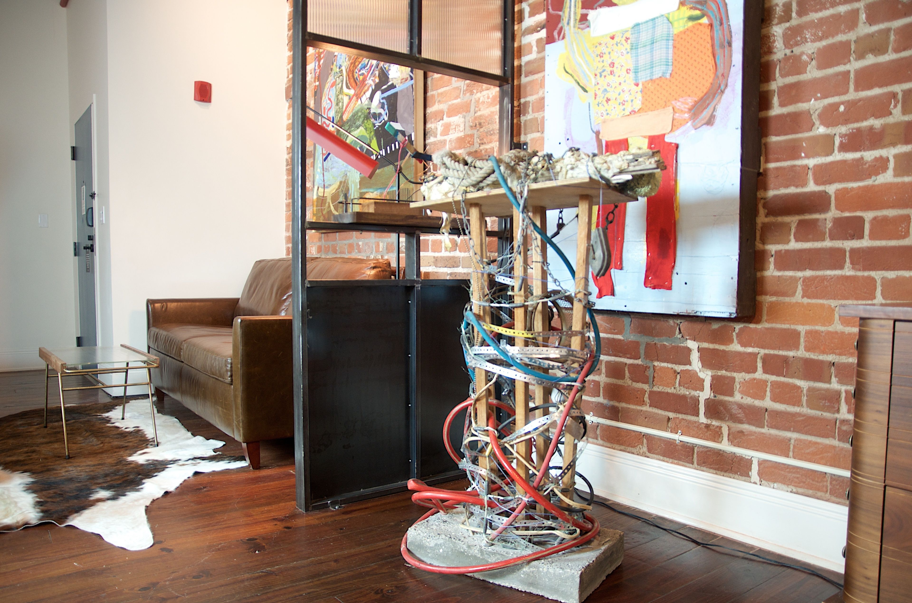 A room with a brick wall, a brown leather sofa, a glass coffee table, and a large abstract painting.
