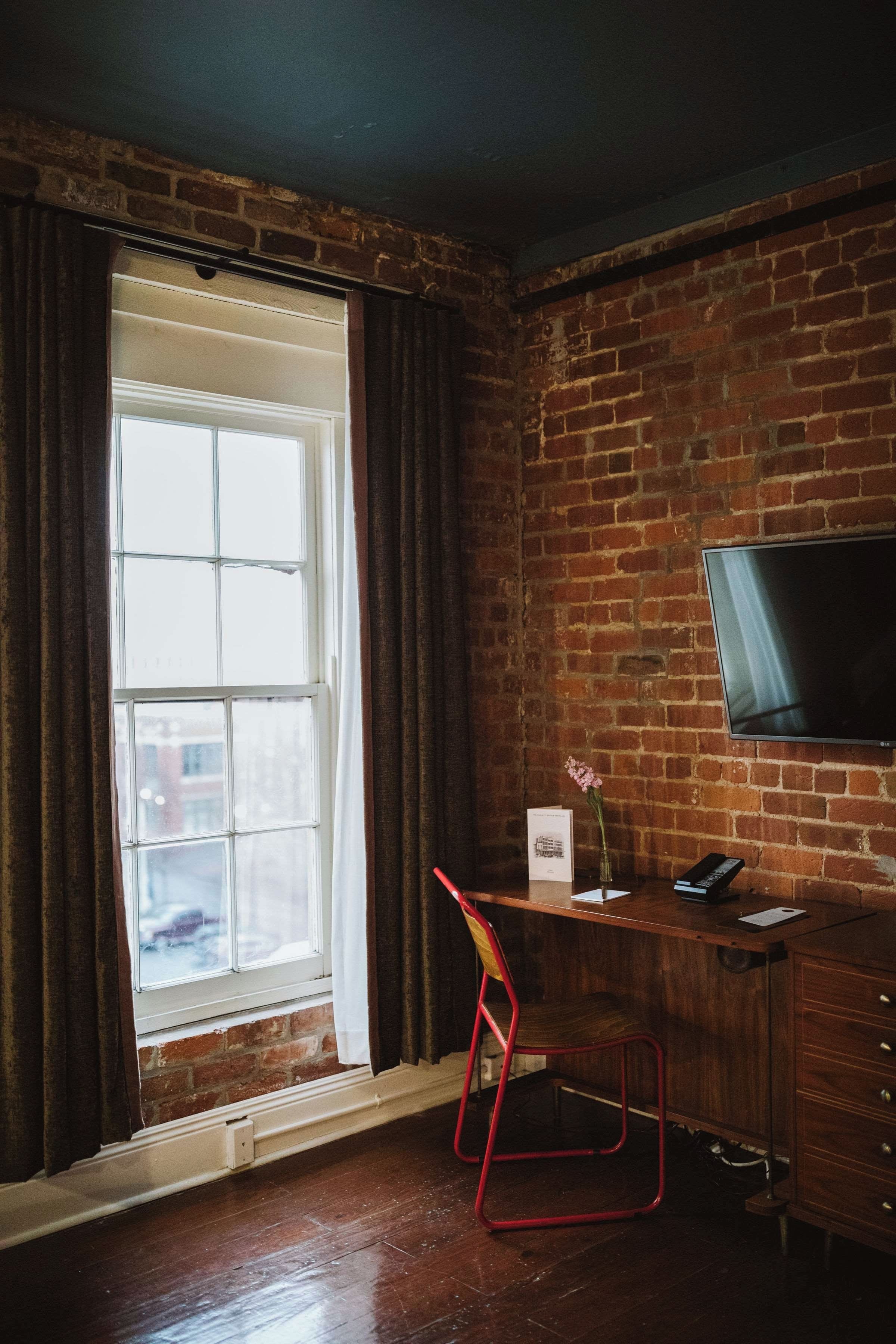 A room with a brick wall, a window with curtains, a desk with a chair, a flat-screen TV, and a vase with flowers.