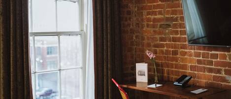 A room with a brick wall, a window with curtains, a desk with a chair, a flat-screen TV, and a vase with flowers.