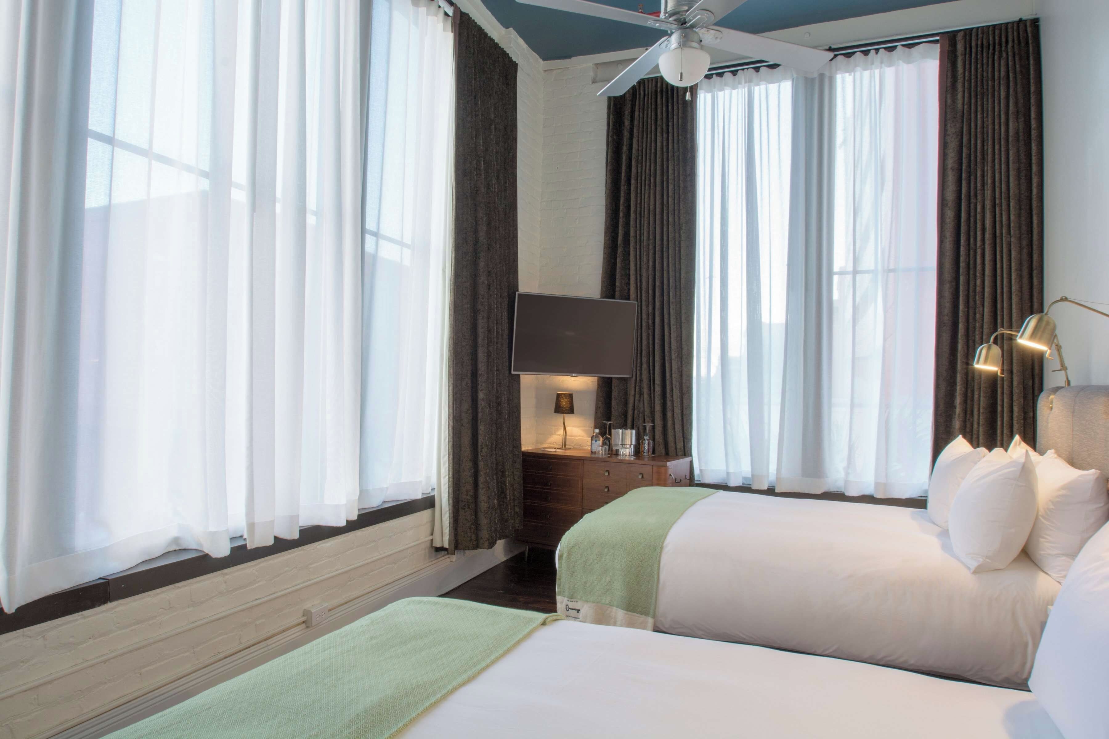 A hotel room with two beds, a flat-screen TV, a desk with a lamp, and a ceiling fan.