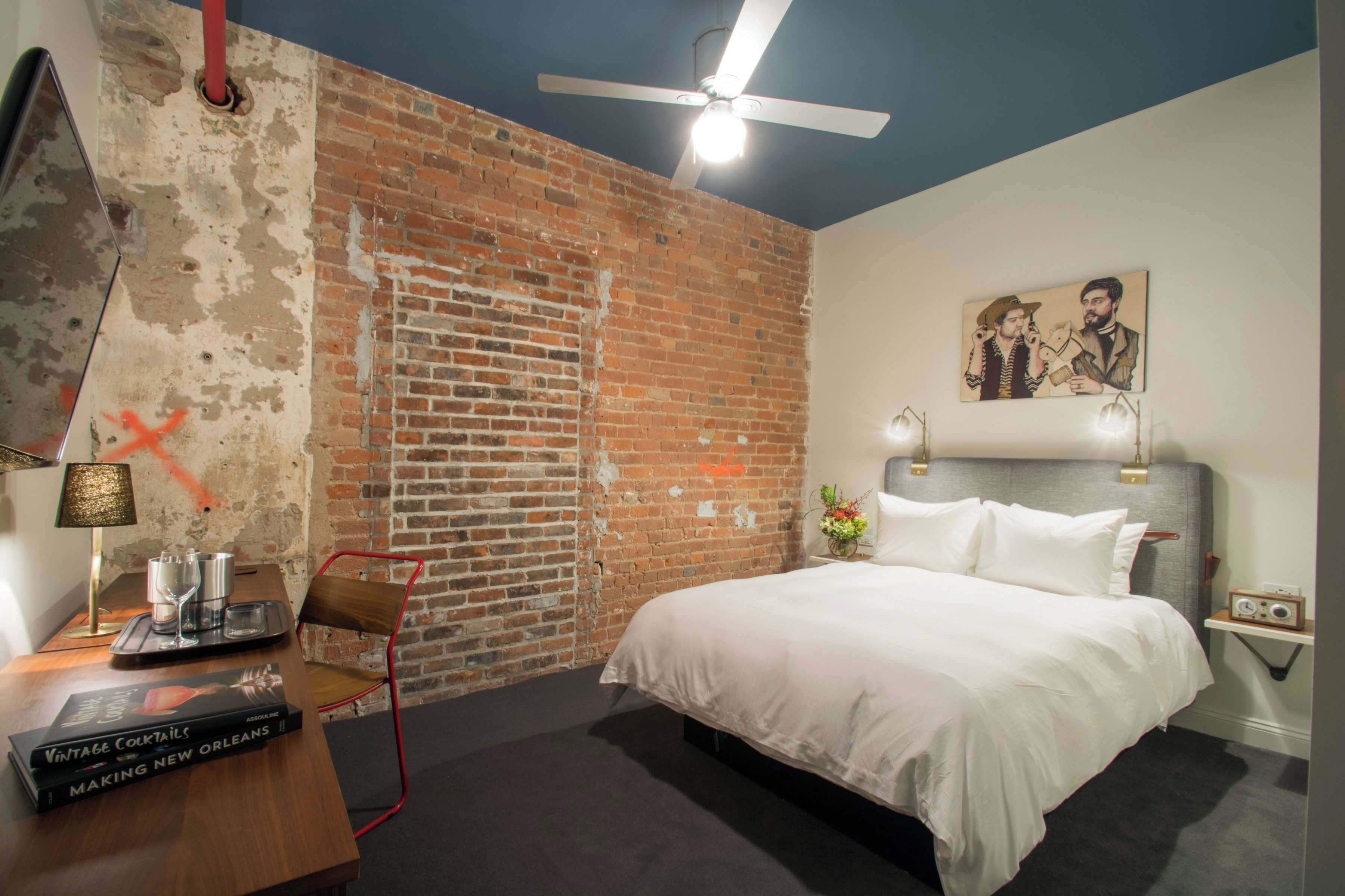 A hotel room with a brick wall, a bed, a desk with a lamp, a chair, and a painting on the wall.