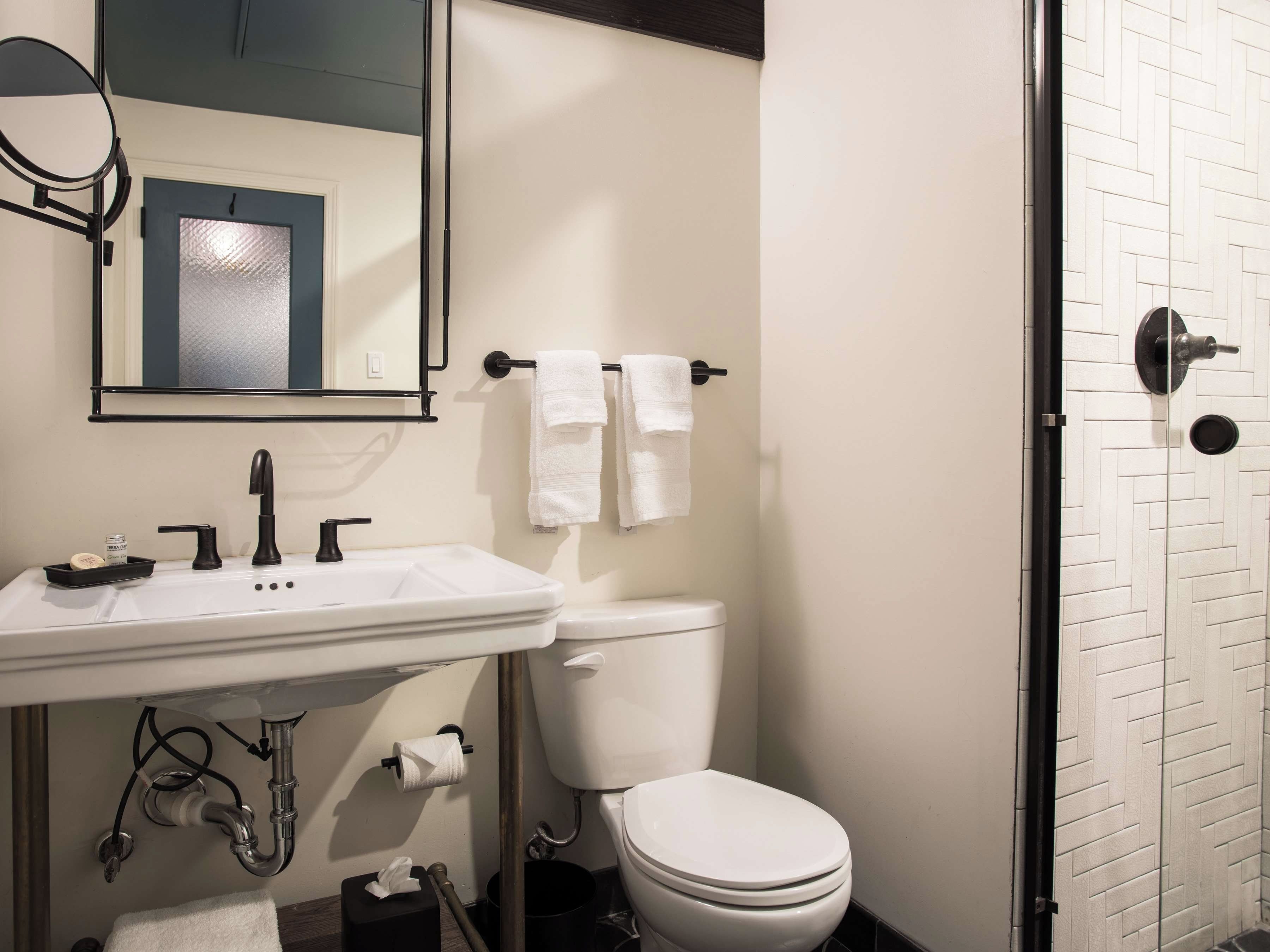 Un baño con un lavabo blanco, grifo negro y un inodoro. Hay un espejo sobre el lavabo y un perchero con dos toallas.