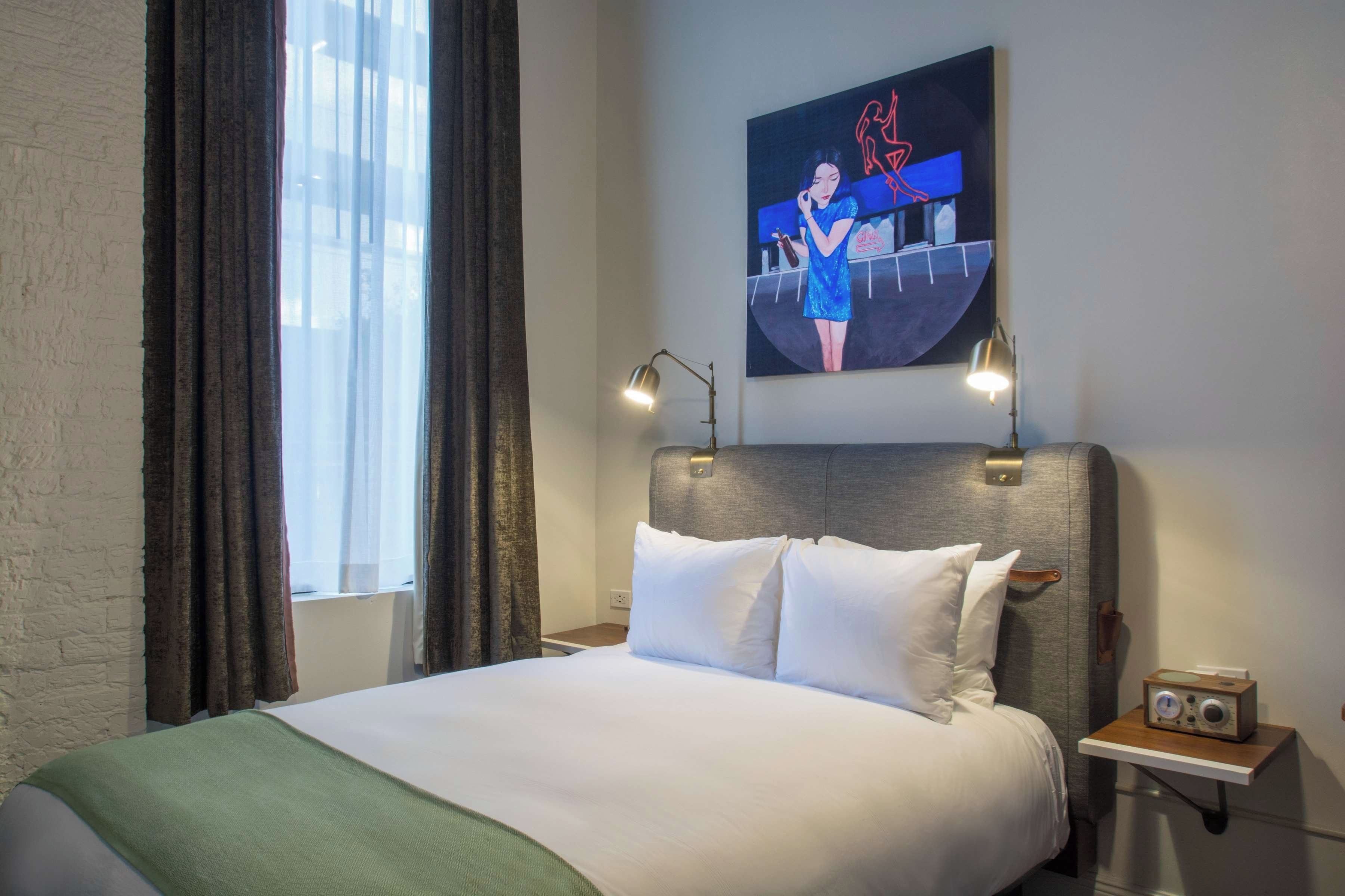 A hotel room with a bed, a nightstand with a radio, a painting on the wall, and a window with curtains.