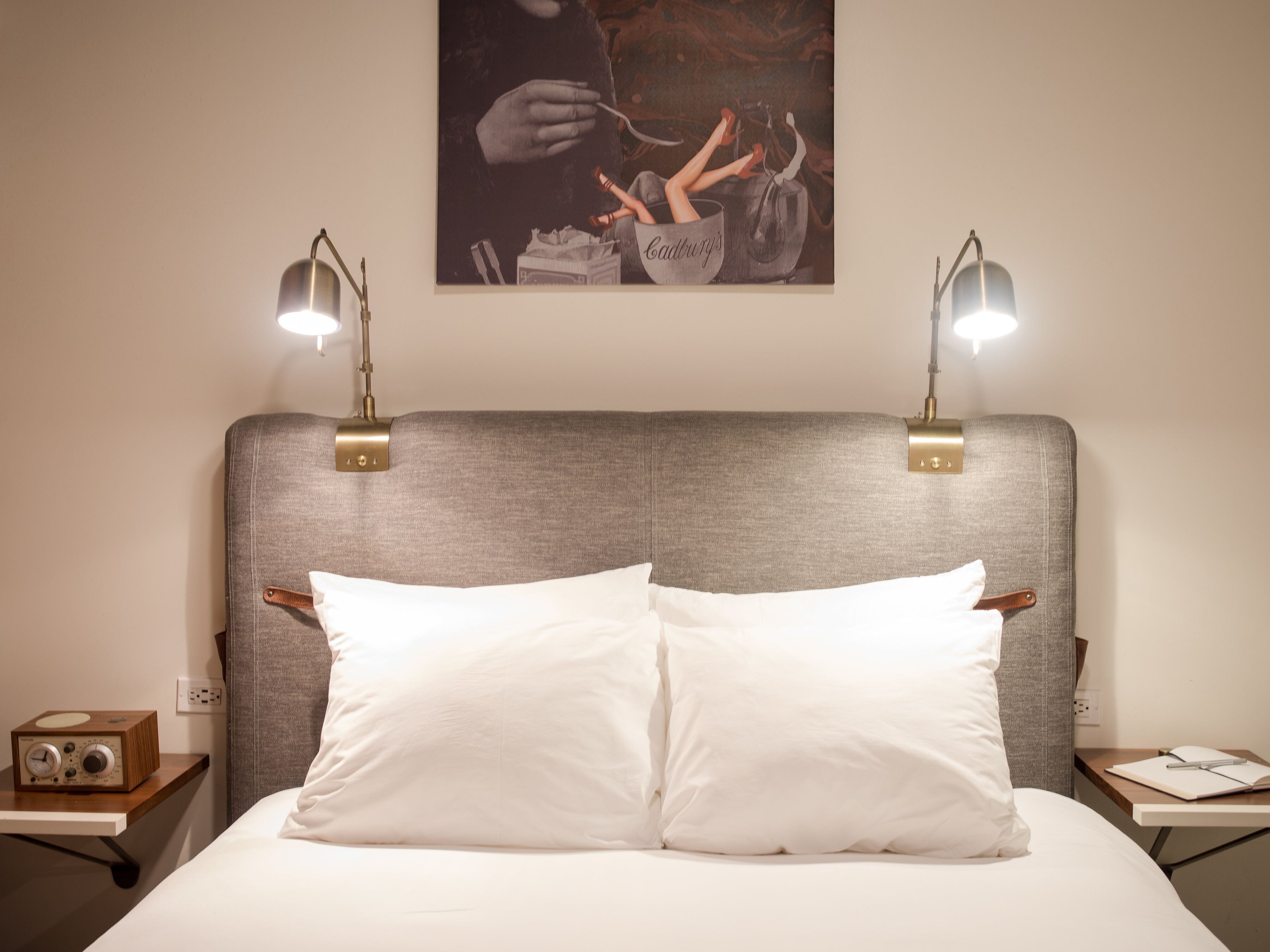 A neatly made bed with white pillows and a grey headboard. There are two bedside tables, each with a lamp and a small radio. A framed picture hangs on the wall above the bed.
