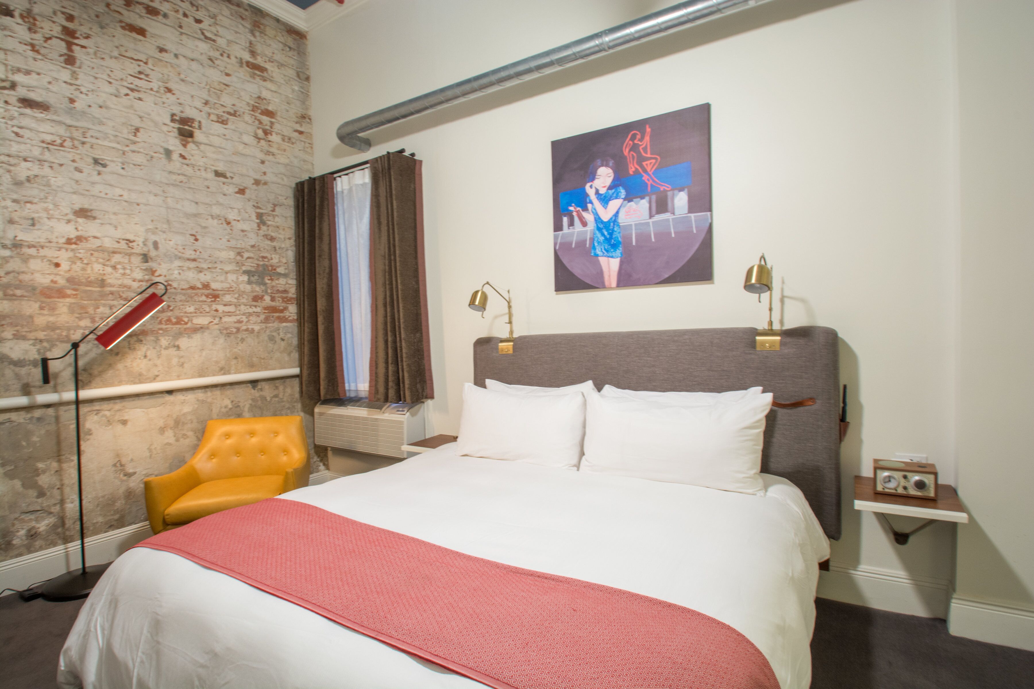 A hotel room with a brick wall, a bed with white and red bedding, a yellow armchair, a painting on the wall, and a bedside table with a radio.