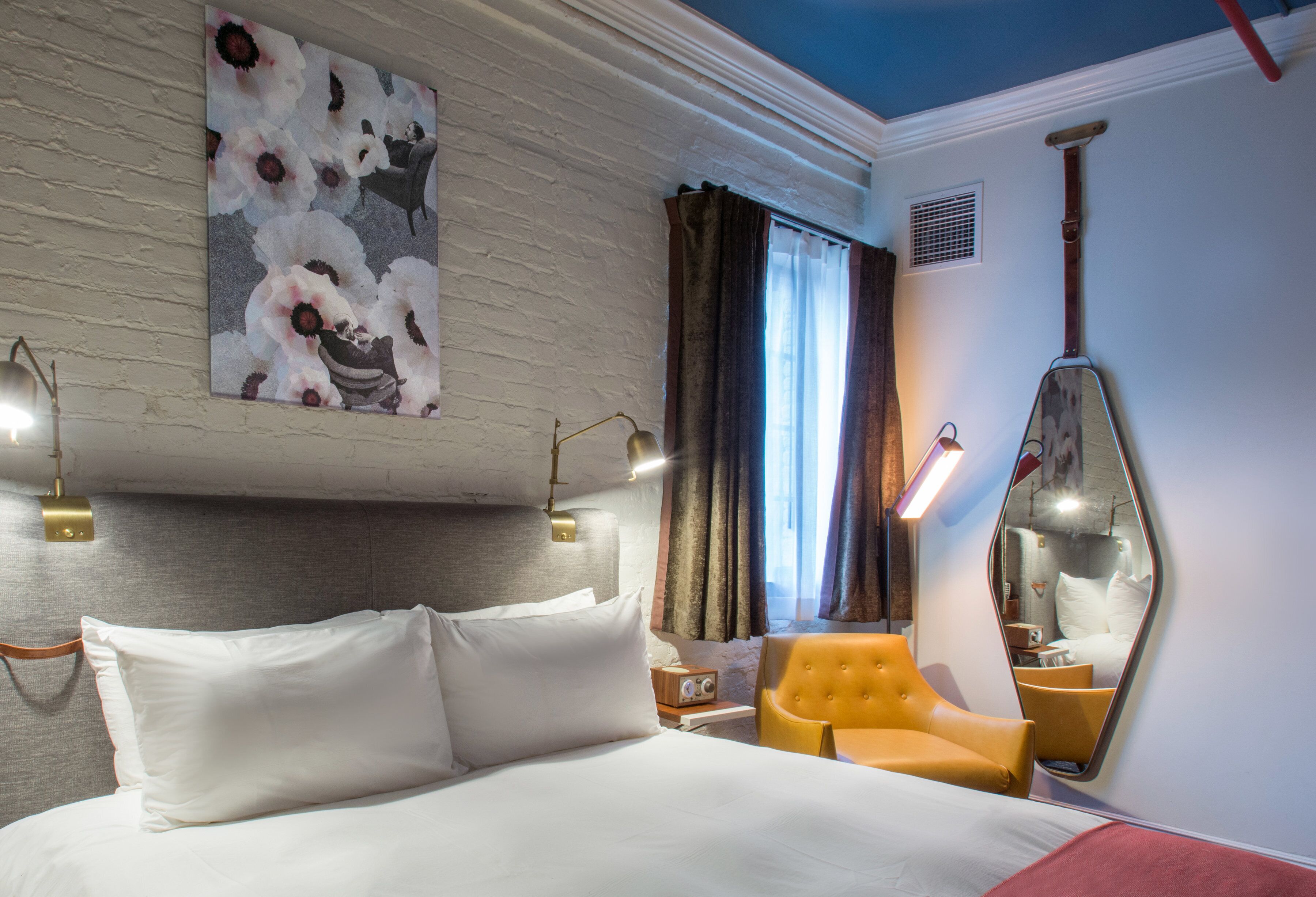 A hotel room with a large bed, a yellow armchair, a wall-mounted mirror, and a painting of flowers.