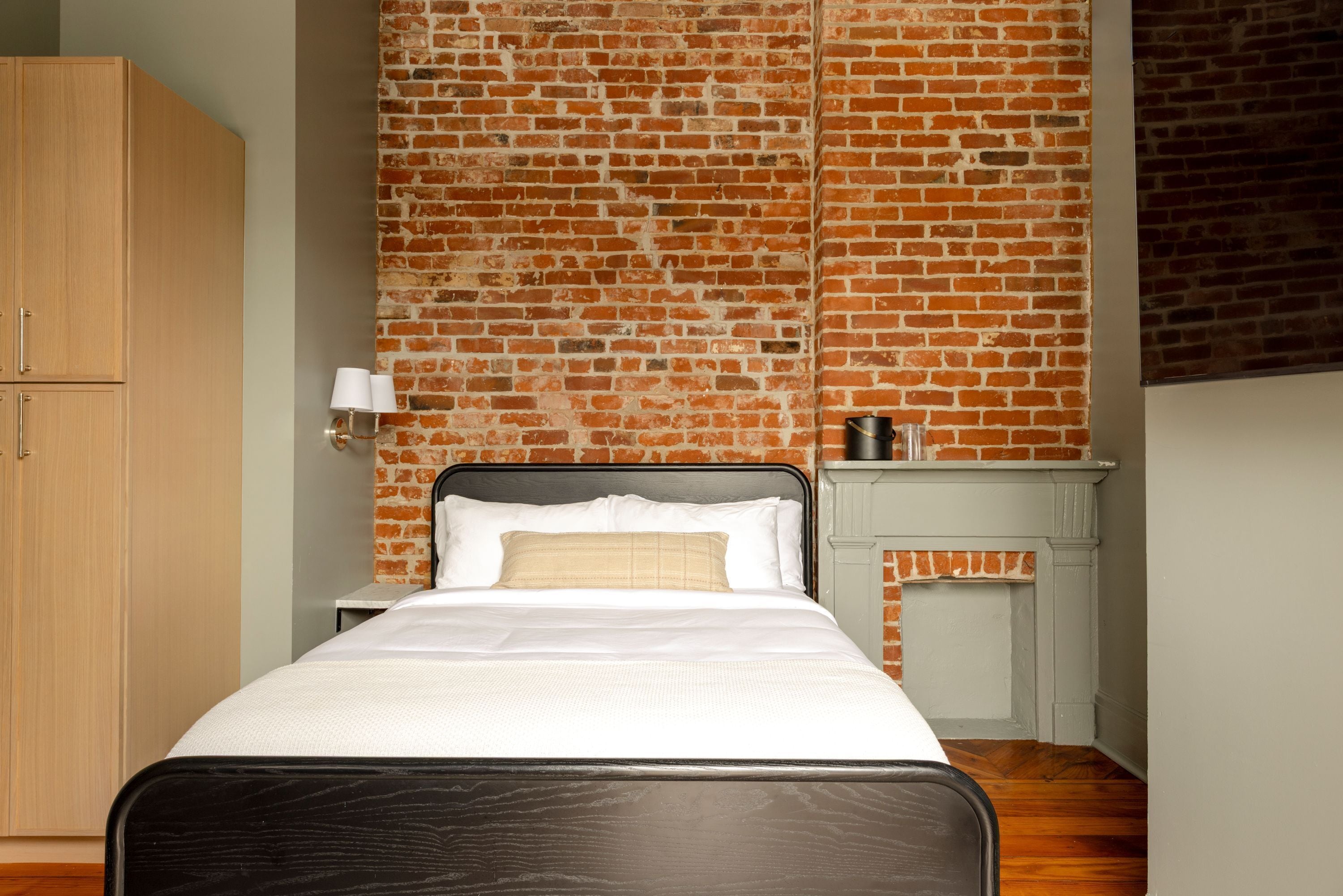 Un dormitorio con pared de ladrillo, una cama con ropa de cama blanca, una mesita de noche con lámpara y una pequeña chimenea.