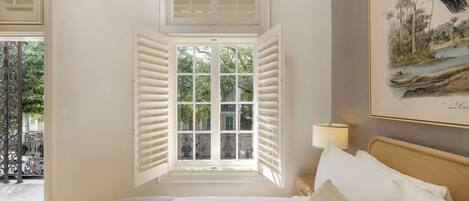 A bedroom with a large bed, a window with shutters, a wall-mounted lamp, and a framed picture.