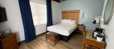 Una habitación de hotel con piso de madera, una cama con sábanas blancas, un escritorio con lámpara, una mesita, una silla, un televisor y una ventana con cortinas azules.
