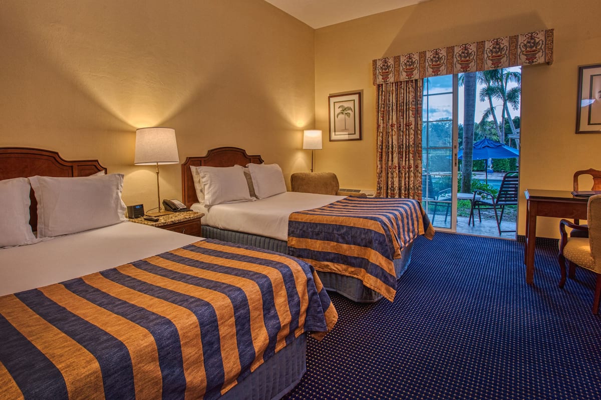 A hotel room with two beds, a desk, and a view of palm trees through the window.