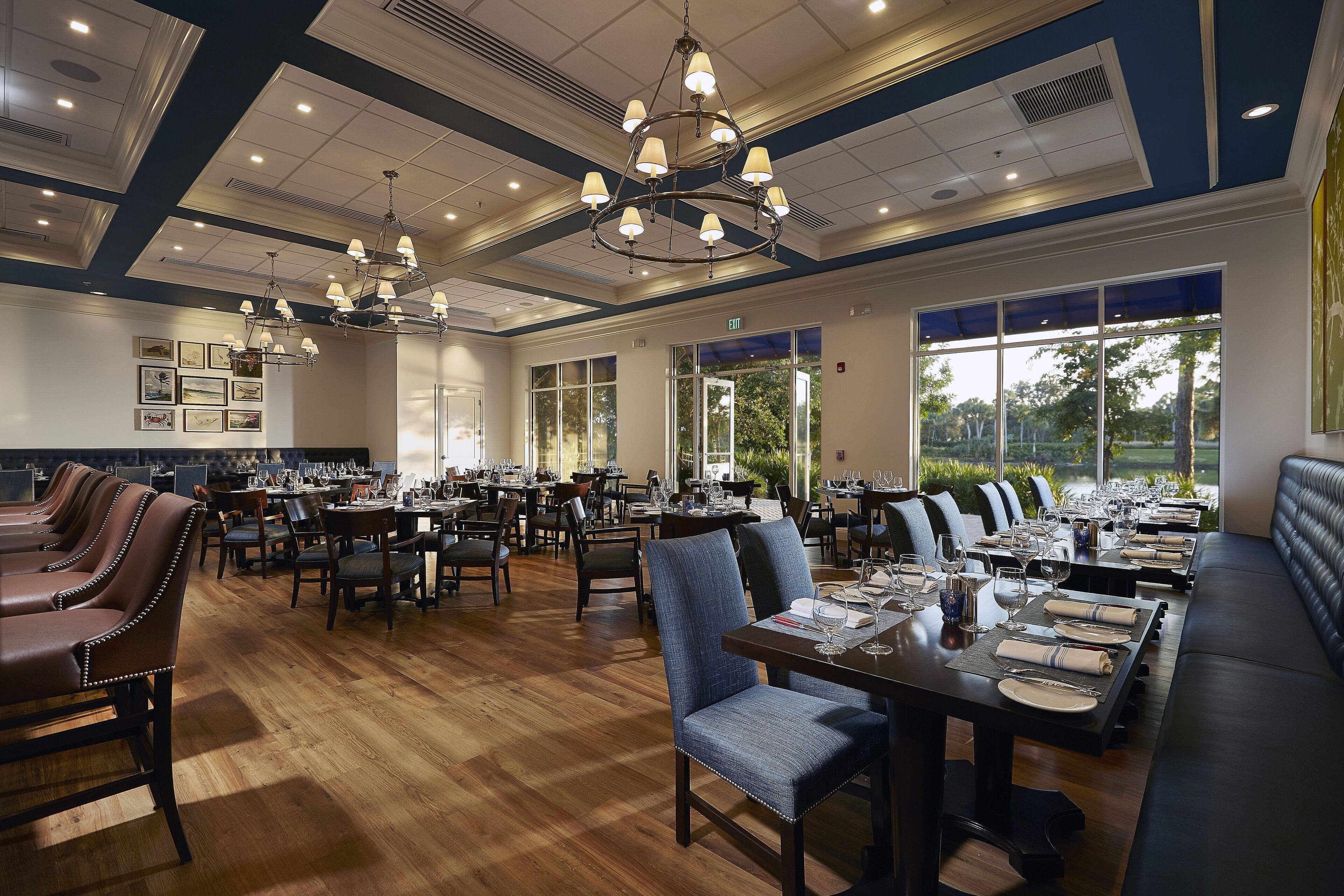 A spacious dining area with multiple tables and chairs, large windows with a view of greenery, and a chandelier with multiple lights.