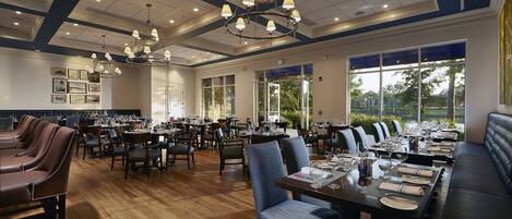 A spacious dining area with multiple tables and chairs, large windows with a view of greenery, and a chandelier with multiple lights.