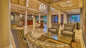 A spacious lobby with high ceilings, large columns, and a chandelier. There are multiple seating areas with patterned chairs and a glass coffee table.