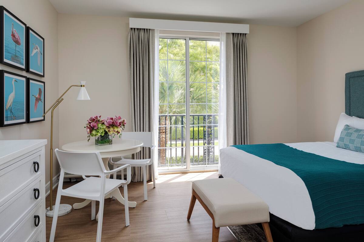 Una habitación de hotel con cama, escritorio con silla, una mesita con flores, cómoda y balcón con cortinas.
