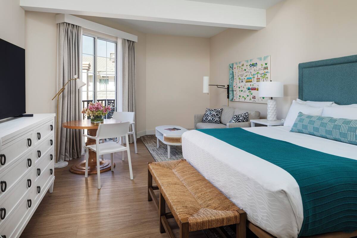 Una habitación de hotel con cama, escritorio, silla, banco, televisor y ventana con cortinas.