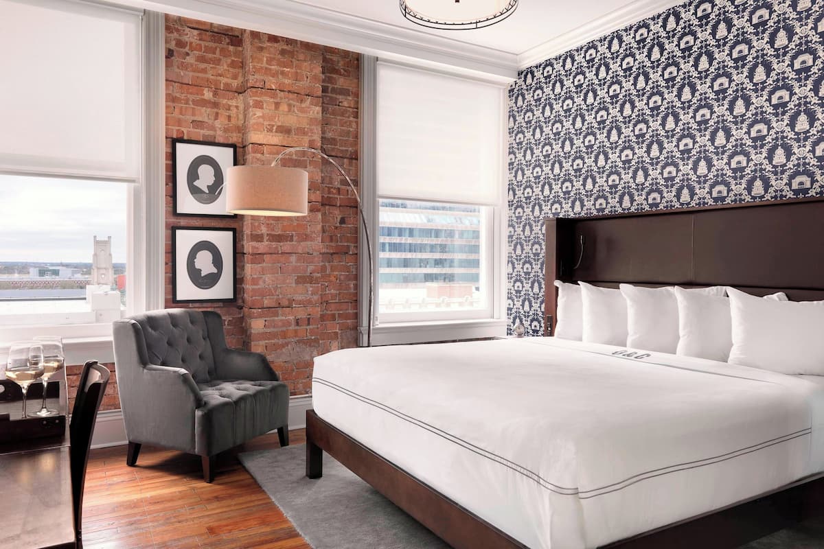 Une chambre d’hôtel moderne avec un grand lit, un fauteuil gris, un mur en briques et un papier peint à motifs.