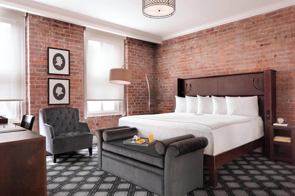 Une chambre d’hôtel avec un mur en briques, un lit recouvert de draps blancs, un fauteuil gris, une petite table sur laquelle se trouve une assiette de tranches d’orange et un verre de jus d’orange, et une lampe sur pied.