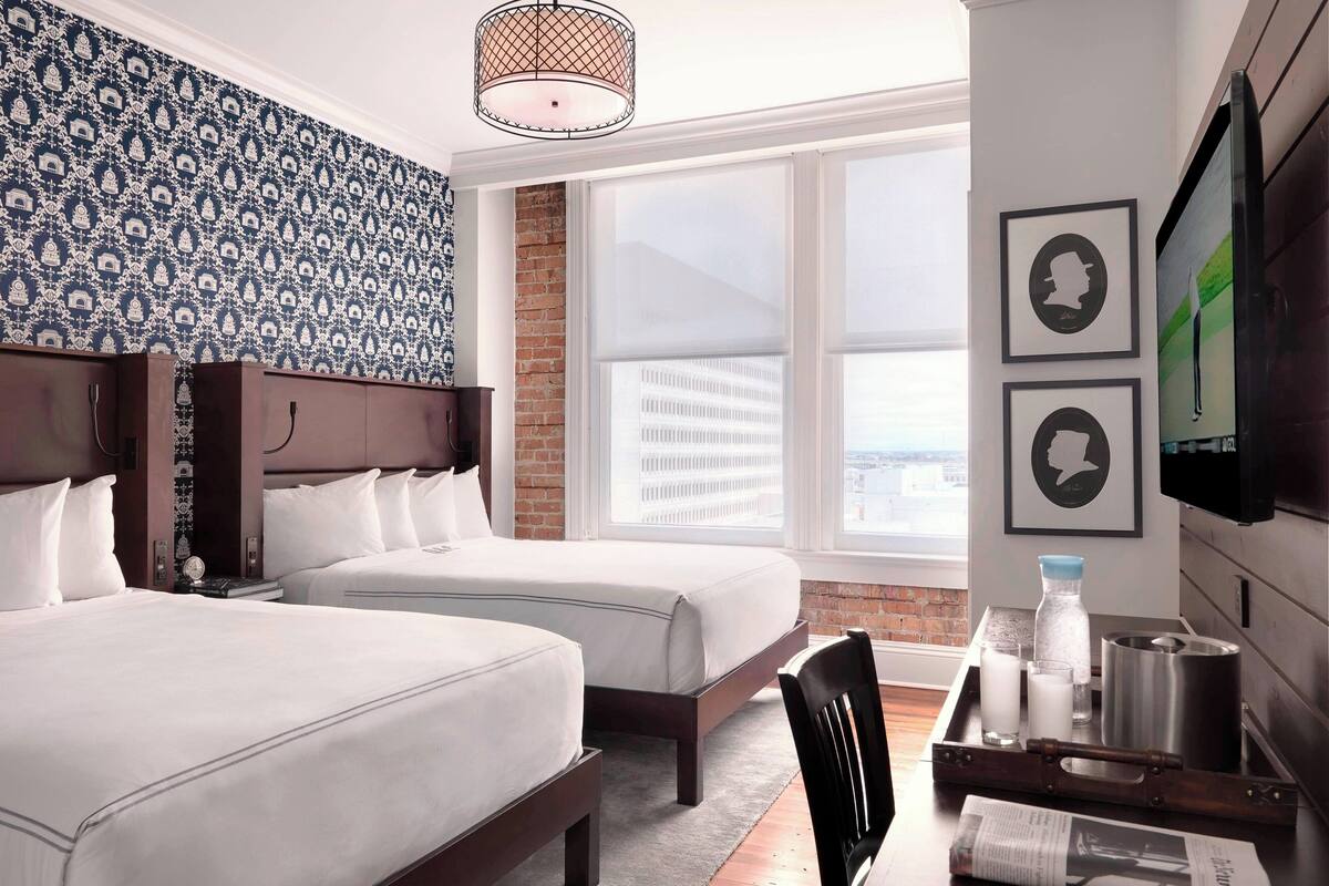 Une chambre d’hôtel avec deux lits, un mur en briques, une grande fenêtre et un téléviseur fixé au mur.