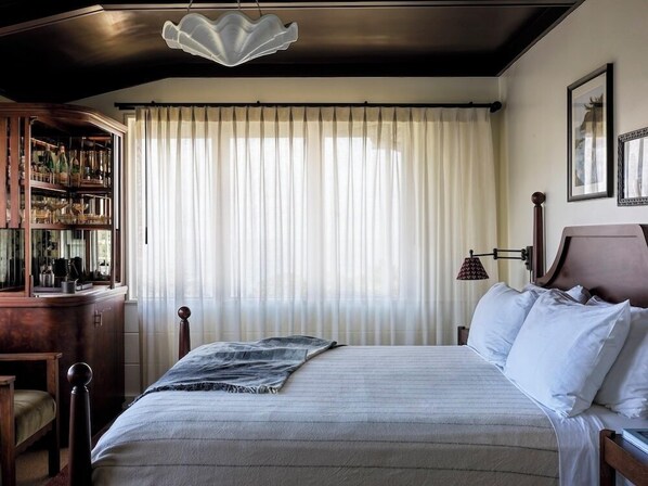 A bedroom with a bed, a wooden headboard, a bedside table, a lamp, a window with sheer curtains, and a decorative ceiling light.