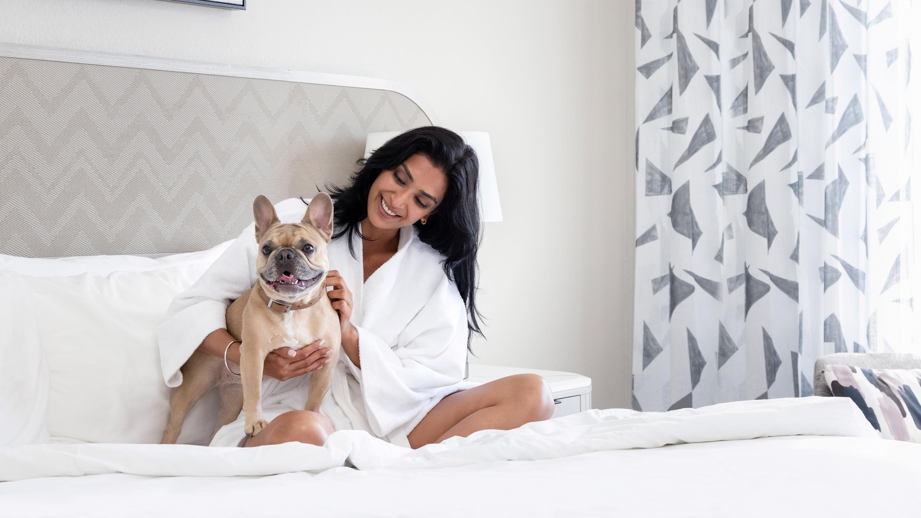 A person sitting on a bed with a dog, holding a remote control, in a room with a patterned curtain and a plain wall.