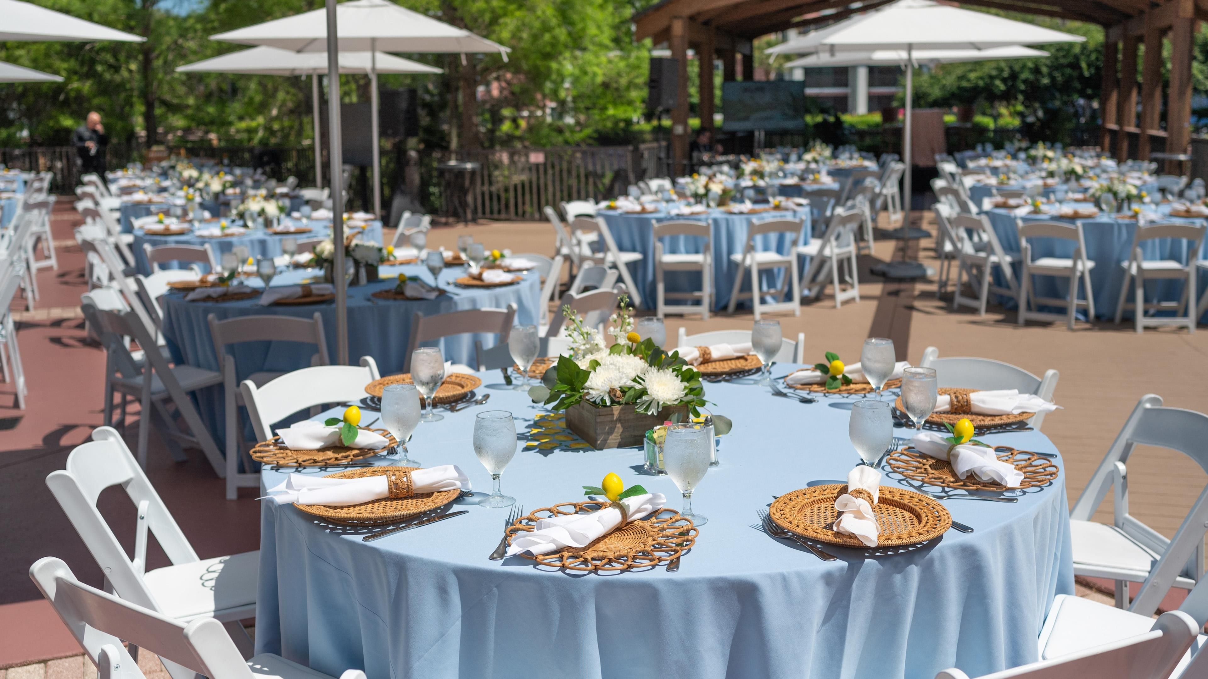 Un área de comedor al aire libre con mesas redondas cubiertas con manteles azules, sillas blancas y sombrillas.