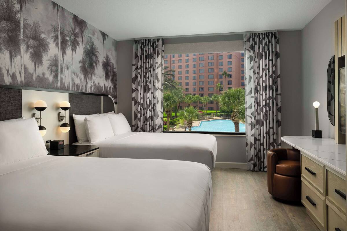 A hotel room with two beds, a large window with a view of a swimming pool and buildings, and a modern design.