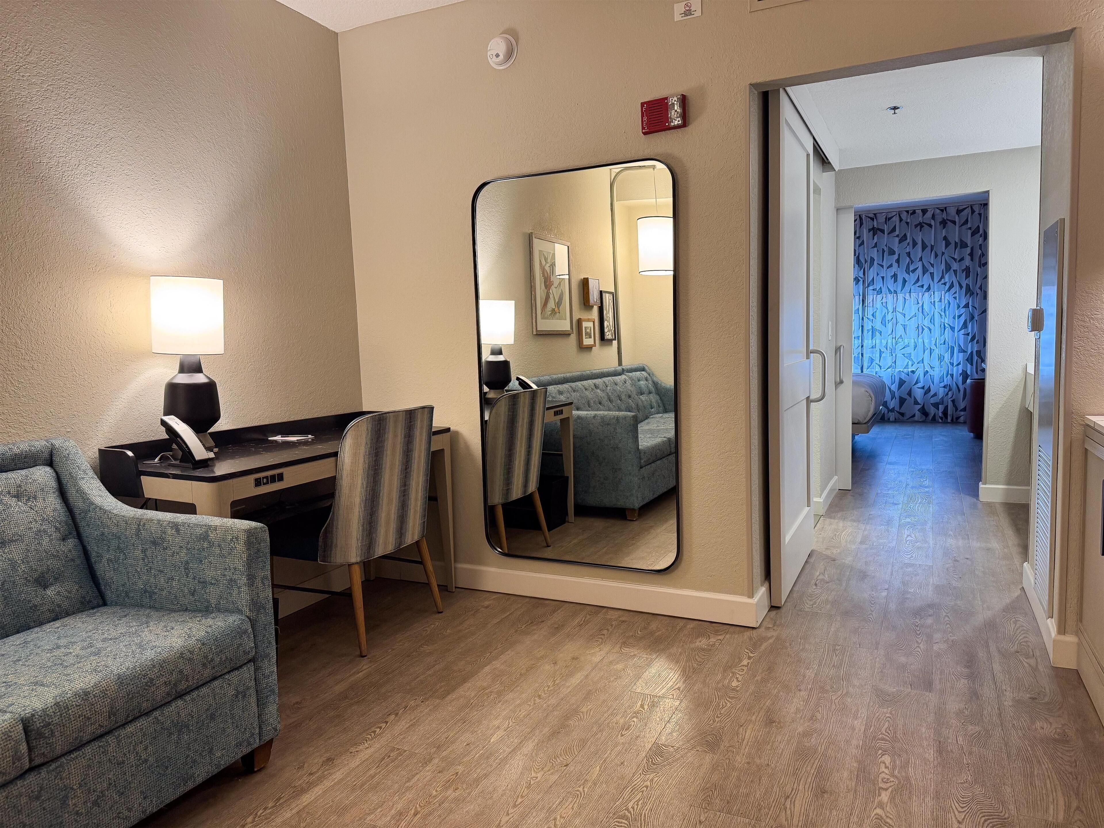 A hotel room with a desk, chair, sofa, and a large mirror.