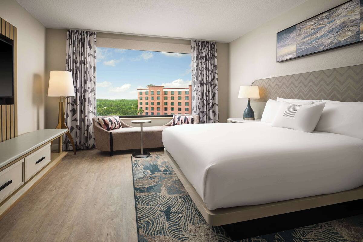 A hotel room with a large bed, a desk, a chair, a small table, and a view of a building through the window.